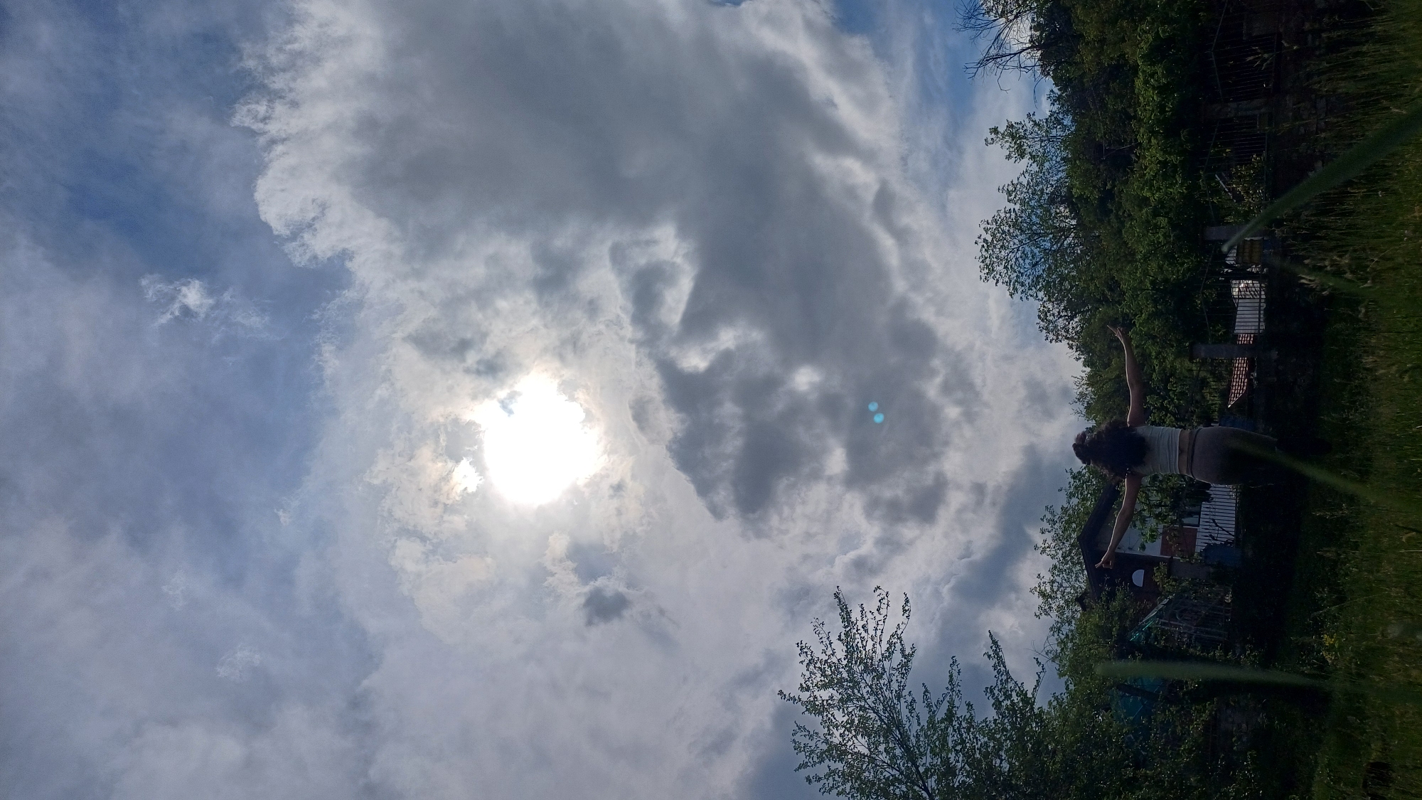 A wide-angle, low-angle shot angled upward reveals a cloudy sky with the sun partially obscured. In the bottom right, a person stands with arms outstretched against a background of trees and structures. Green vegetation and possibly a sprinkler line are visible in the foreground. The location is Gorna Verenitsa, Bulgaria, as deduced from geographical information.