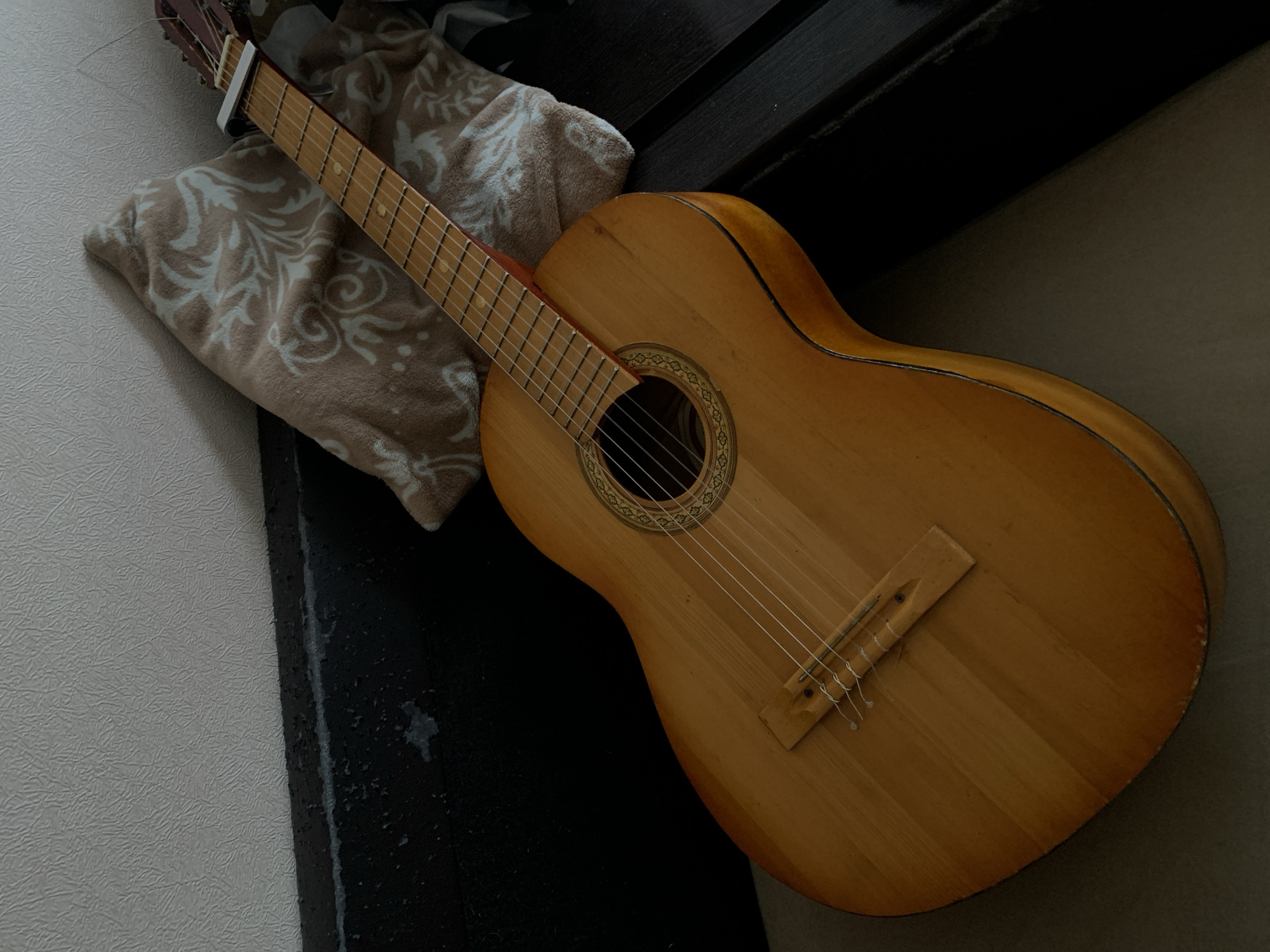 The image shows a classical guitar resting on a patterned pillow. The guitar has a light wood finish and six strings. It is leaning against a wall or corner. The environment appears to be indoors, with a dark surface adjacent to the pillow. The city and specific location are unknown.