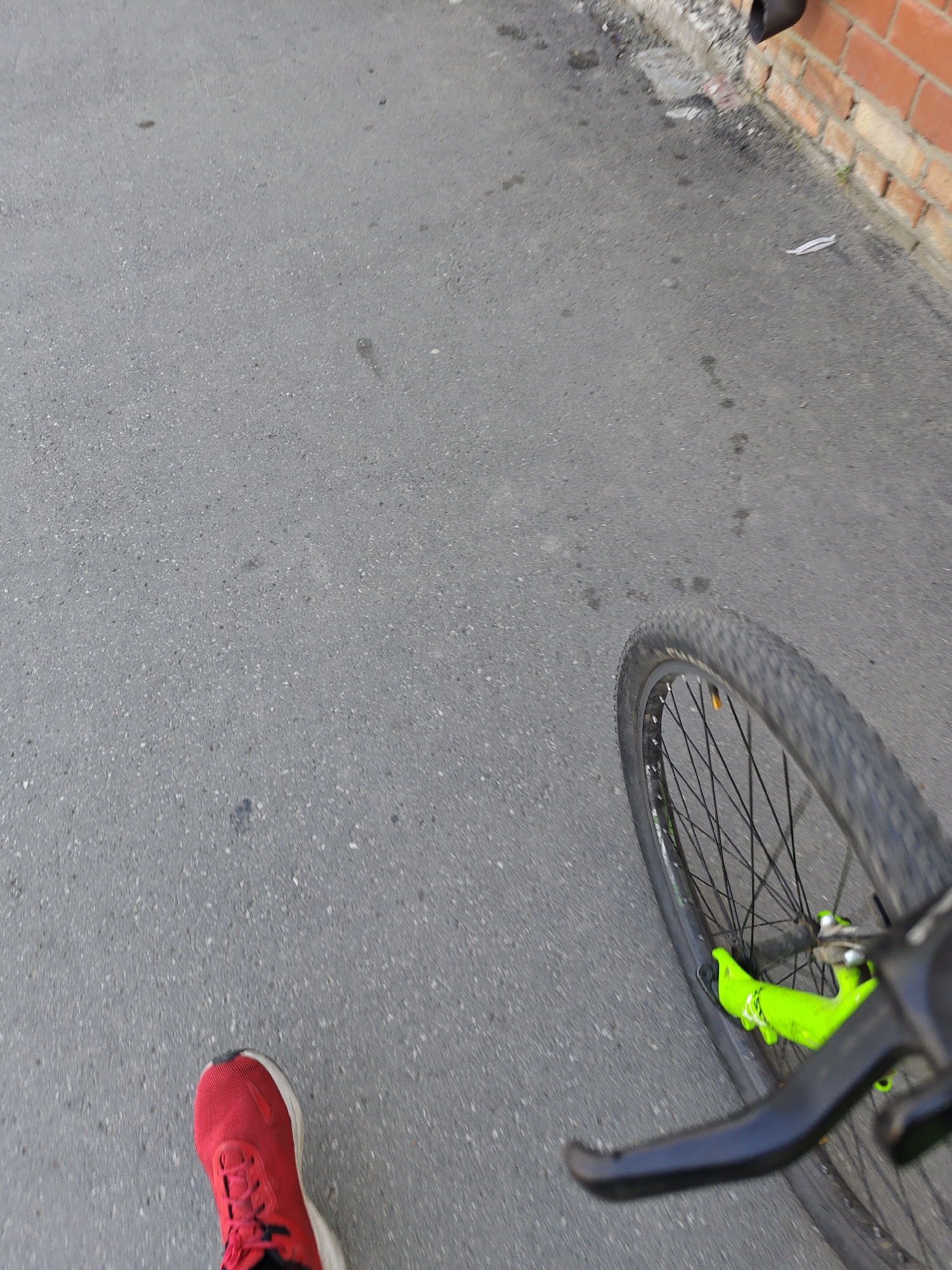 A first-person perspective shot shows a red shoe on an asphalt surface next to a bicycle wheel. The bicycle has a black tire and rim with spokes, and a bright green frame. Part of the bicycle's brake lever is visible in the foreground. In the background, a brick wall runs along the edge of the asphalt. The overall scene suggests movement, potentially someone riding or pushing a bicycle.