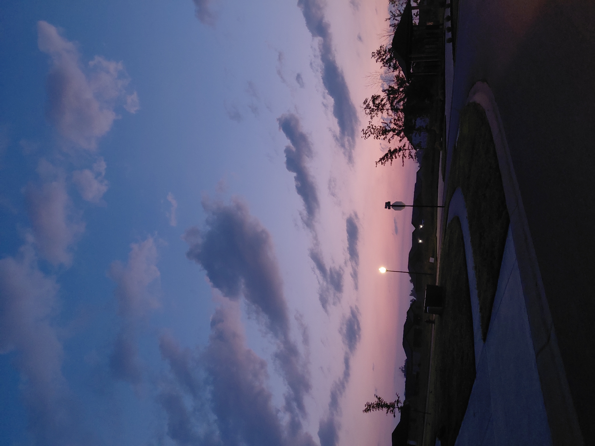 The image depicts a suburban street scene at dusk in Richmond, United States. The sky is a gradient of deep blue at the top transitioning to soft pink and lavender hues near the horizon, dotted with scattered clouds. A single street lamp illuminates the area with a warm glow, casting light on a sidewalk and a curved asphalt road. Several houses with dark silhouettes are visible in the background, along with mature trees that are also silhouetted against the twilight sky. A small gazebo-like structure is present on the right side of the image. The scene conveys a sense of quiet and transition as day turns into night.