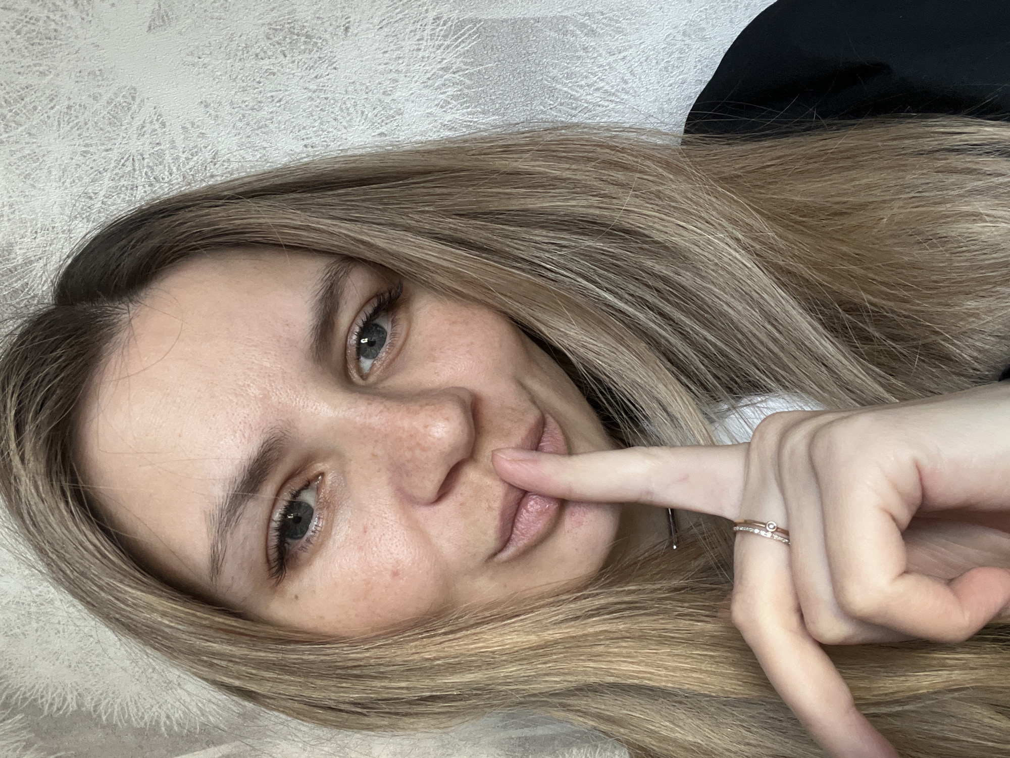 A female subject, appearing to be a young adult, is depicted in a close-up indoor portrait. She has long, blonde hair and blue eyes, with visible freckles across her nose and cheeks. Her right index finger is pressed vertically against her lips, indicating a "shush" gesture, while she looks directly into the camera. She is wearing a dark-colored garment, possibly a jacket or top, with a white collar visible beneath it on the right side of the frame. Two rings are worn on her right ring finger: a plain gold-toned band and another gold-toned band featuring small clear stones and a central circular embellishment. The background is a light-colored wall with textured wallpaper displaying subtle, abstract linear patterns. The scene is situated indoors, with the presumed location being the city of Chaikovsky, Russia.