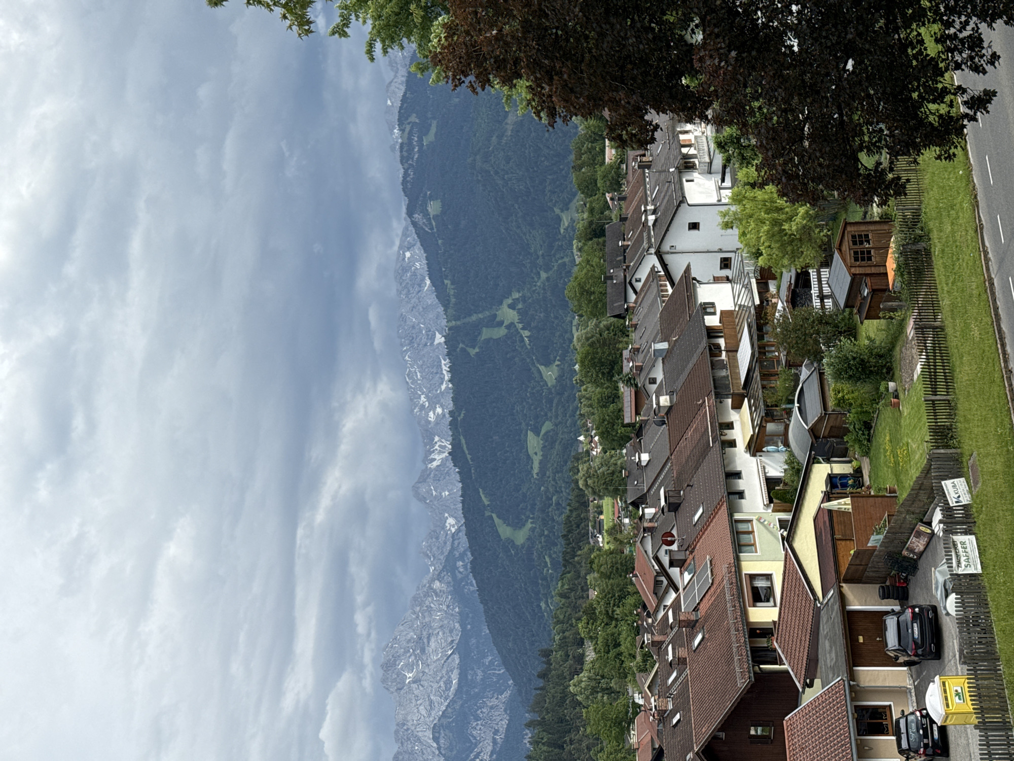 The image captures a panoramic view of a residential area in Garmisch-Partenkirchen, Germany, set against a dramatic backdrop of snow-capped mountains under a cloudy sky. The foreground is dominated by a cluster of houses with reddish-brown tiled roofs, typical of Bavarian architecture. Several of these homes feature solar panels, indicating a modern adaptation to traditional design. In the mid-ground, more houses are visible, interspersed with lush green trees and smaller structures, possibly sheds or garages. Two black cars are parked in front of one of the houses, beside a yellow portable toilet and some stacked tires. A wooden fence delineates a grassy yard, where a small wooden shed stands. Further in the distance, the scene transitions to dense, dark green forests that climb the slopes of the mountains. The mountains themselves are rugged and imposing, with visible patches of snow on their peaks and ski slopes etched into their sides, suggesting they are part of the Alps. The sky is overcast with heavy, grey clouds, creating a somewhat somber mood but also highlighting the texture of the mountainous terrain. The overall impression is one of a peaceful, established community nestled within a majestic natural landscape.