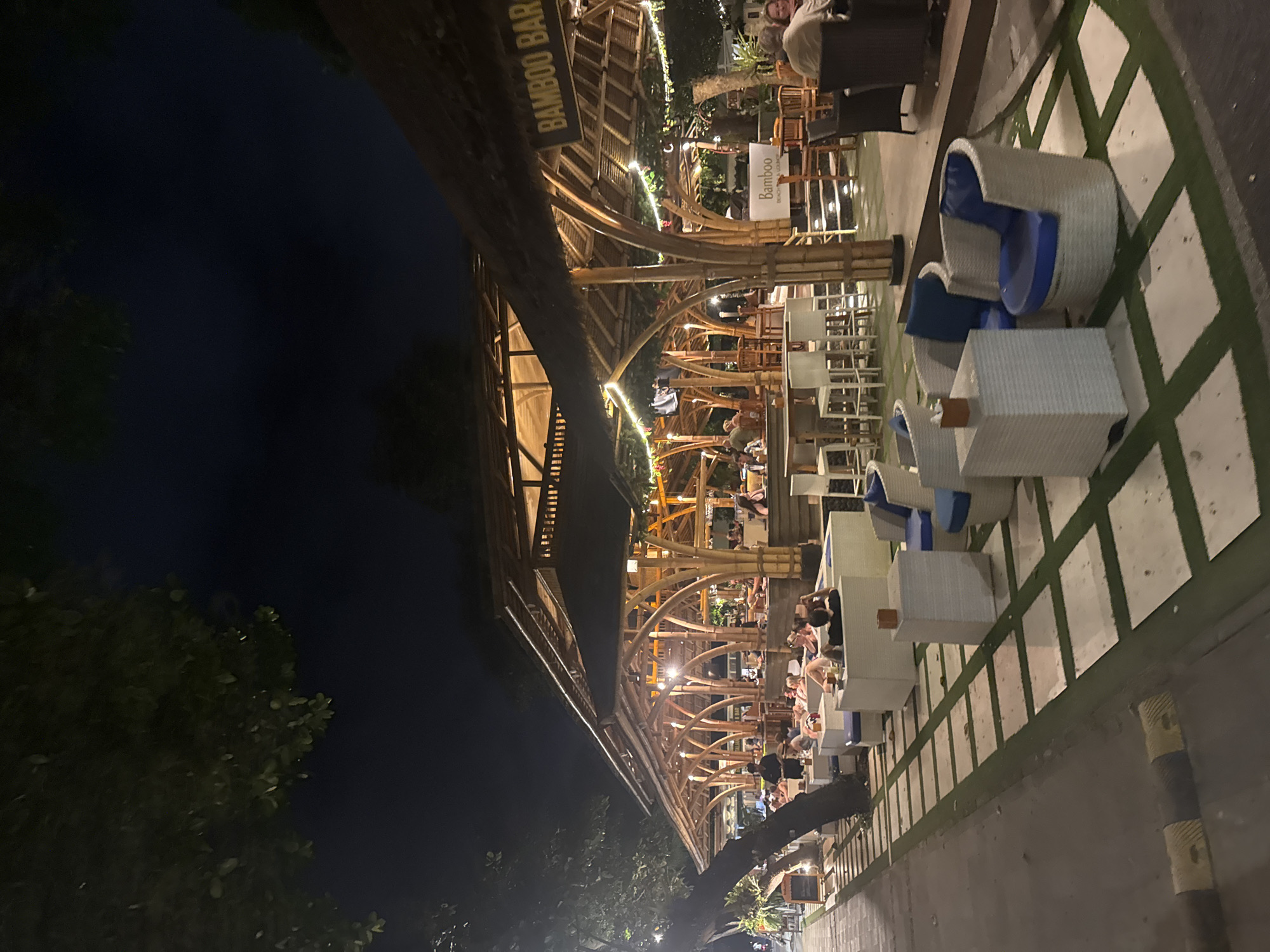 The image shows an outdoor nighttime scene at "Bamboo Bar" in Denpasar, Indonesia. The bar has a bamboo structure with lights strung throughout. People are seated at tables and chairs, appearing to be eating and drinking. The seating area is adjacent to a paved area, and there is greenery to the side. The sky is dark, suggesting it is night.