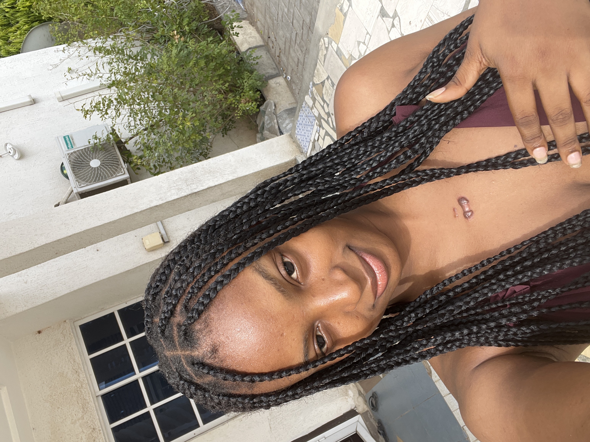 A young adult female is depicted in a close-up, outdoor setting within the city of Lokoja, Nigeria. She has a dark skin tone and wears long, dark box braids, partially obscuring her shoulders. Her face is directed towards the camera with a slight smile. A prominent, raised, light-colored keloid scar is visible on her upper central chest. She is wearing a dark red or maroon garment, partially visible. Her right hand, with manicured nails, rests on her upper chest, near the neckline of her garment.
The background consists of a light-colored, stucco-textured building wall. To the left, a white-framed window with six dark panes is visible. To the right, a "Hisense" branded outdoor air conditioning unit is mounted on the wall, above green leafy shrubbery. A circular white light fixture is positioned higher on the wall, and a white vertical pipe runs adjacent to the air conditioner. The ground beneath the subject and extending into the background is paved with irregular, multi-colored (light grey, white, brown) tiles, transitioning to a grey concrete curb and further paved surfaces. The scene is illuminated by natural daylight.