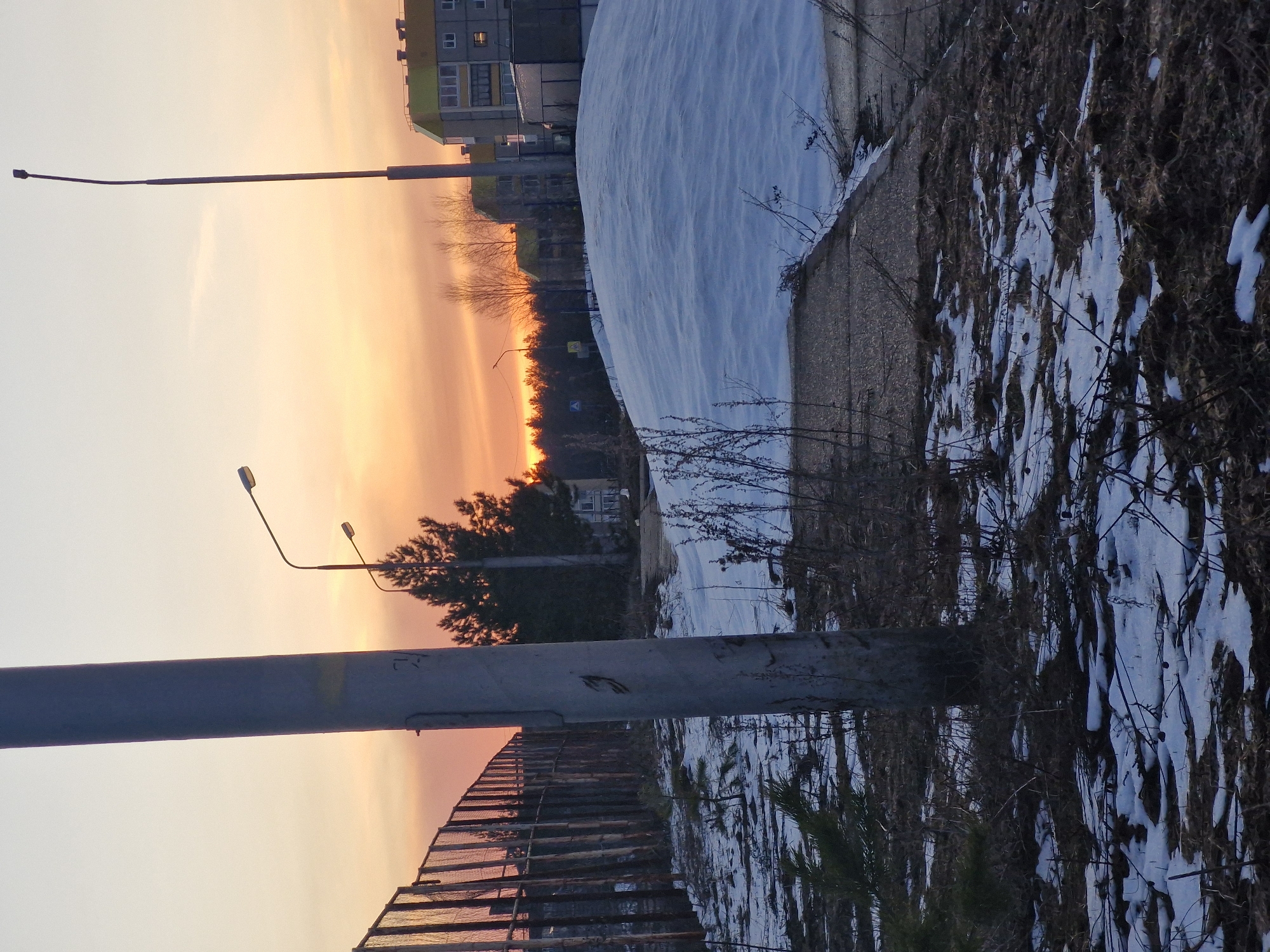 The image shows a cityscape during sunset, possibly in Markovskii, Russia. A large snowdrift covers the ground near a concrete wall, interspersed with dry vegetation and patches of melting snow. Two streetlights are visible, with the glow of the sunset illuminating the sky behind trees and buildings. A metal fence is also present in the foreground.