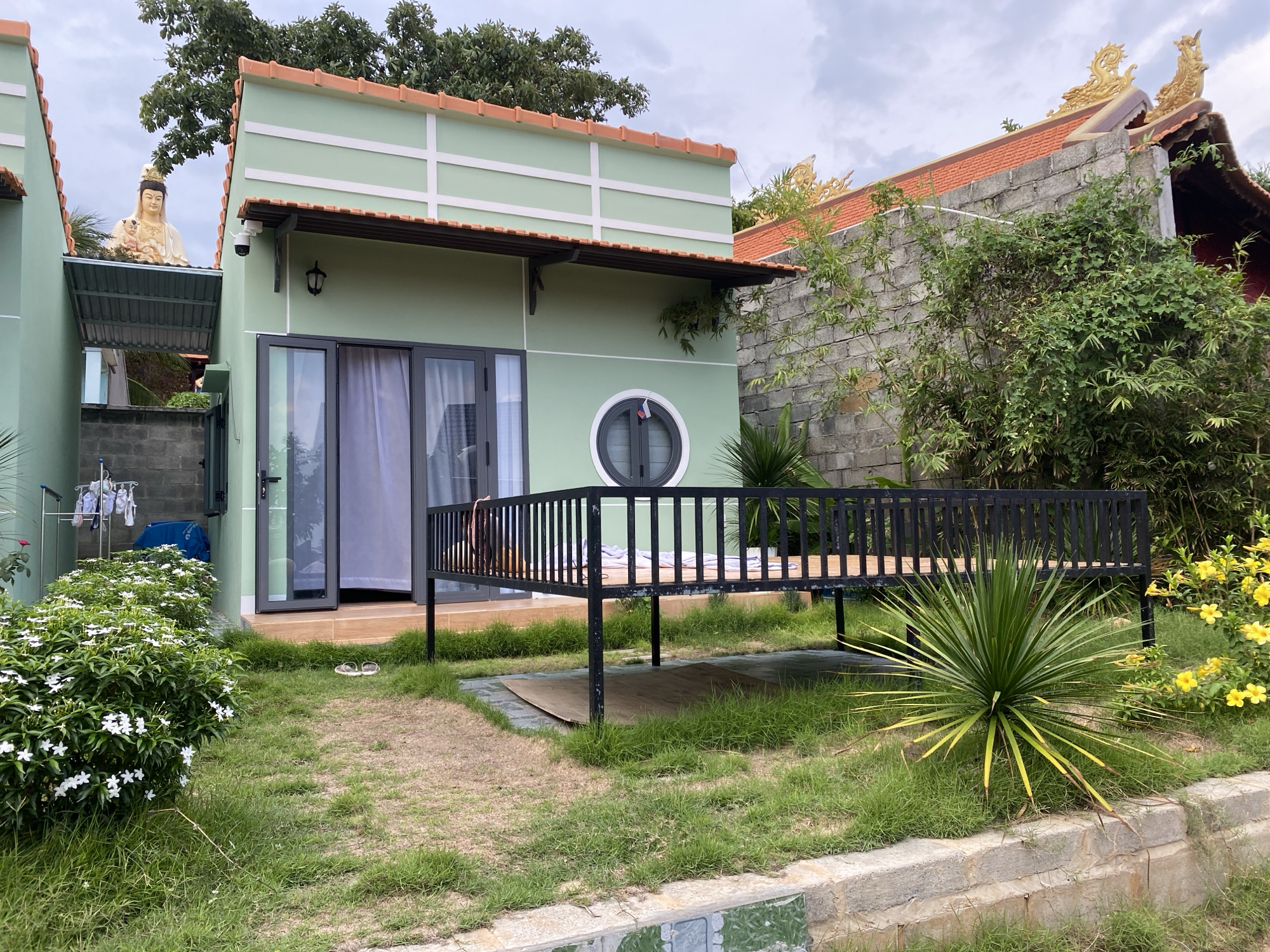 This image shows a house in Mũi Né, Vietnam, with a light green facade, a terracotta-colored roof, and white trim. The house has a sliding glass door, a round window, and a black metal deck. In front of the house, there is a lawn with various plants, including a yucca plant with long, pointed leaves, a bush with white flowers, and yellow flowers. A statue of a woman holding a child is on the roof of the adjacent building to the left. Another building, with more ornamentation and a terracotta-colored roof, is visible behind the house to the right. The sky is overcast.