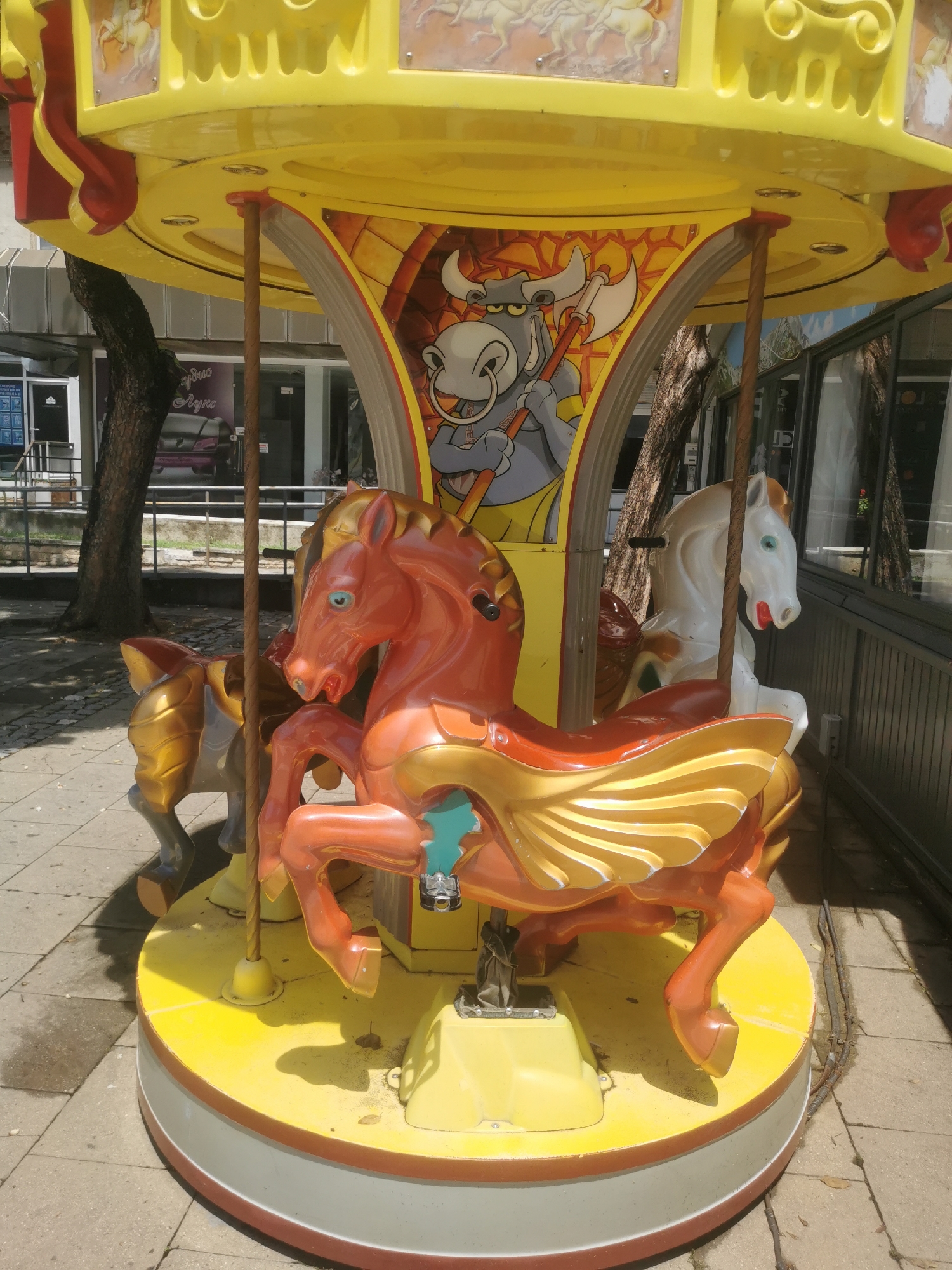 This media file captures a brightly colored, outdoor children's carousel, likely situated on a paved sidewalk or public square in Blagoevgrad, Bulgaria. The carousel features a predominantly yellow structure with red and gold accents.
The main subject is an orange and gold winged horse, positioned prominently in the foreground. It has bright blue eyes. Behind it, parts of another orange and gold horse are visible, and to the right, a white horse with a red mouth and light brown mane can be seen. All horses are mounted on poles, ready for riding. The yellow circular base of the carousel appears somewhat worn and dusty.
The carousel's canopy is also yellow and features decorative panels. A prominent central panel depicts a cartoonish, grey bull-like creature with large horns and rings in its nose, holding a double-headed axe. Other panels around the top show more traditional carousel imagery of horses, though these are less distinct.
The setting is outdoors during a sunny daytime, evidenced by the bright light and clear shadows cast by the carousel and nearby trees. In the background, there are building facades with reflective windows. One storefront on the left side of the image has partially visible text, which appears to be "Studio Lux" (likely "Студио Лукс" in Cyrillic). A large tree with a dark trunk and green foliage stands between the carousel and the buildings. The ground is paved with concrete slabs or pavers. No people are visible, and the carousel appears stationary.
