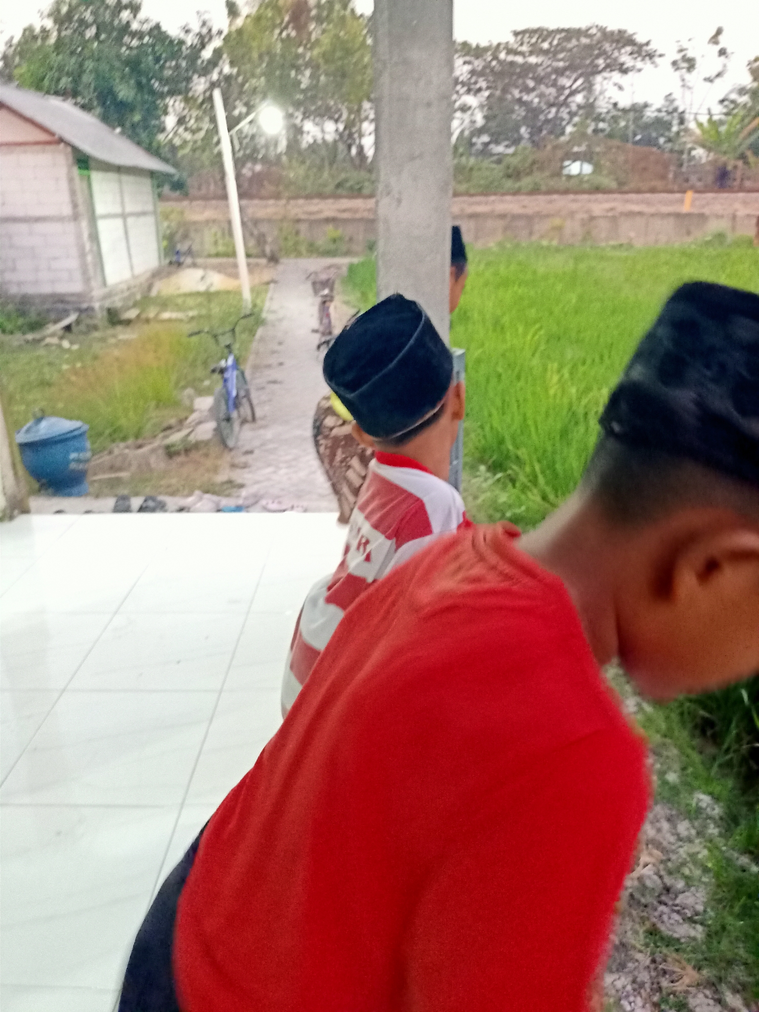 The image depicts an outdoor scene with a young boy in the foreground, facing away from the camera and wearing a red shirt and a black cap. He appears to be standing on a white tiled surface. To his left, another young boy wearing a red and white striped shirt and a black cap is partially visible. In the background, there is a grassy field, a building with a white and light-colored exterior, and a dirt path. There are two bicycles on the path, one blue and the other dark-colored. A blue cylindrical object is also present near the building. A light pole with a lamp is visible between the two boys. The overall environment suggests a rural or semi-rural setting. The location is identified as Kendung, Indonesia.