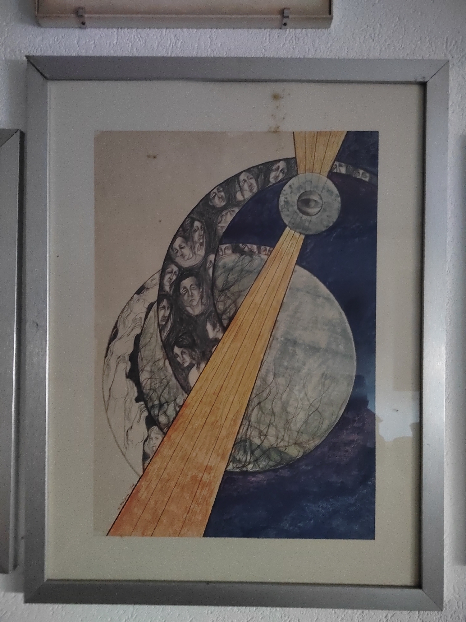 A framed artwork, featuring a surreal composition, hangs on a textured white wall. The artwork itself depicts a swirling vortex of faces and abstract shapes. A prominent element is a large, circular disc with an eye in the center, intersected by a thick, golden beam. The background transitions from a light, cream-colored area to a deep blue, speckled with purples and blues, suggestive of a night sky or cosmic expanse. The overall impression is one of depth and mystery. The location of this artwork is the town of Porto Recanati, Italy, specifically within a private residence.