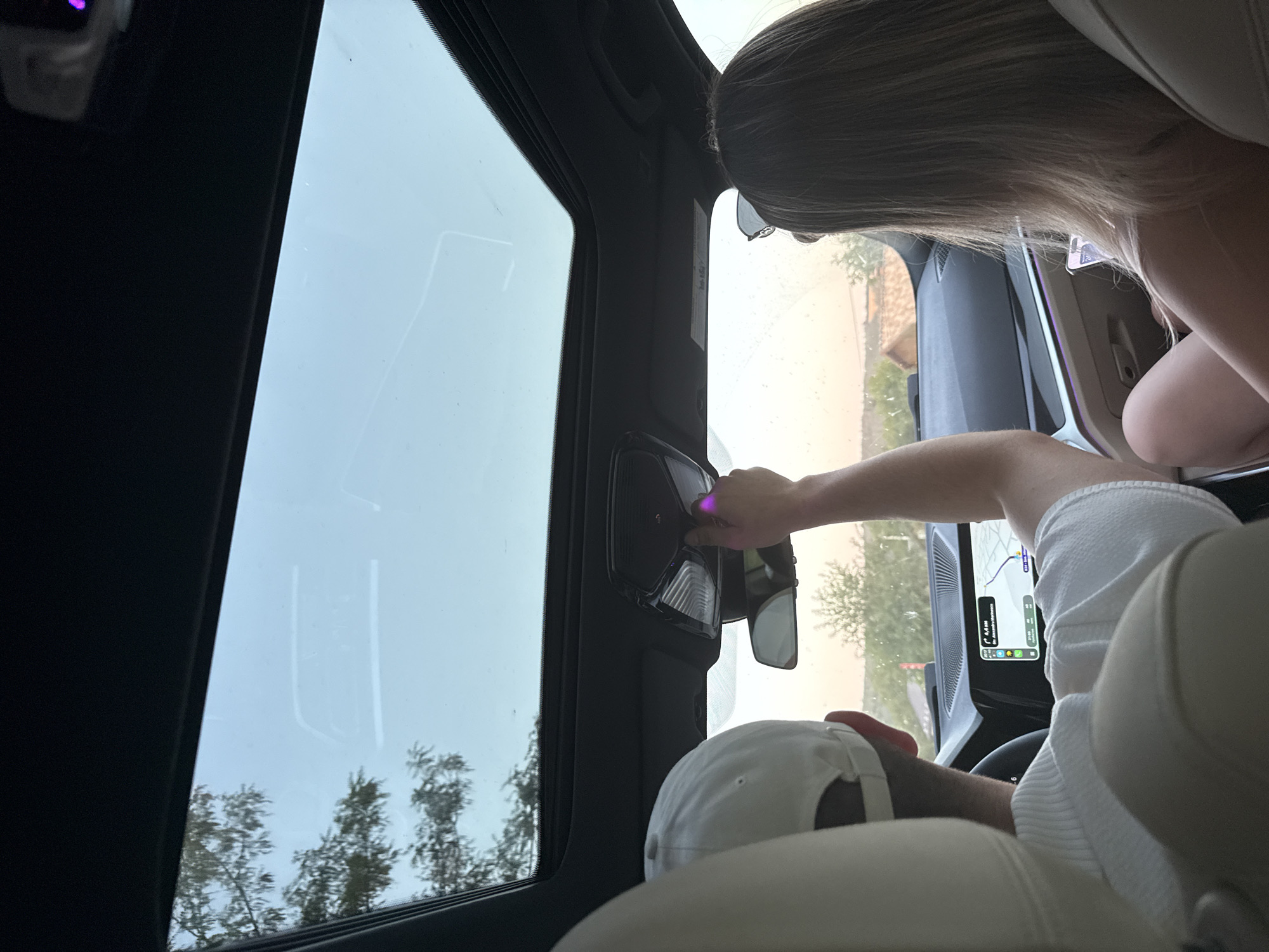 In a car's interior, two individuals are visible from behind, seated in the front. A person on the left wears a white baseball cap and a white shirt. Their right arm is extended upwards, touching the car's ceiling light console, where a faint purple light is emanating. The person on the right has long blonde hair and is wearing a light-colored top and sunglasses on their head. Their left hand rests on their lap, holding a smartphone displaying a screen with numbers.
The car's sunroof is open, revealing a bright, overcast sky. The dashboard shows a navigation screen with a map displaying a route. Outside the car, trees and buildings are visible, suggesting a rural or suburban setting. The ambient light indicates it is either early morning or late afternoon. The specific location is identified as Sărata-Mereșeni, Moldova.