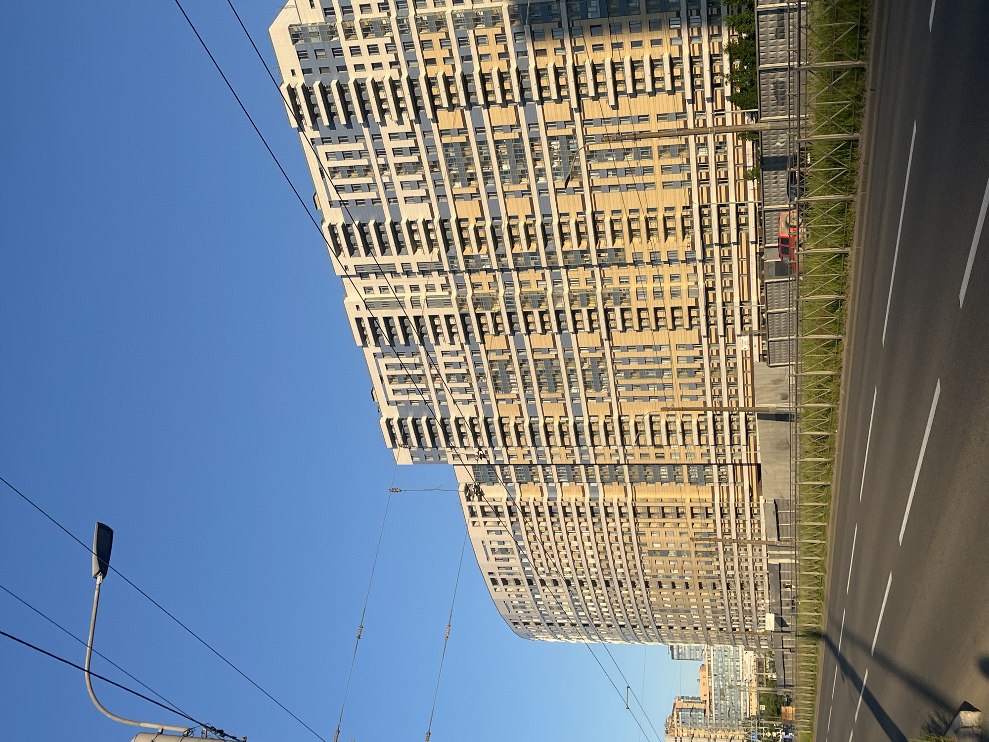 This image depicts an urban street scene on Komendantsky Prospekt in Saint Petersburg, Russia, specifically showcasing the "Legenda Komendantsky" residential complex.
The scene is dominated by a clear, unclouded blue sky, with the sun positioned to the left, casting sharp shadows. In the upper left quadrant, a grey, curved metal streetlight pole extends towards the center, supporting a modern, elongated grey light fixture. Multiple black electrical or tram wires traverse the upper half of the frame at various angles, some attached to insulators.
In the foreground, a multi-lane asphalt road runs horizontally across the bottom of the image. It features white dashed lane markings indicating traffic lanes. To the right of the road, a low, green metal fence with a diamond-patterned mesh runs parallel to the road. Behind this fence, a narrow strip of green grass is visible, followed by a tall, grey concrete barrier or sound wall, textured with recessed square patterns. Behind this barrier, a red SUV-type vehicle and a dark sedan are partially visible.
The midground and background are dominated by the multi-story "Legenda Komendantsky" residential complex. The most prominent structure is a large, curved building on the left, displaying a facade of alternating horizontal bands of light-colored (white/cream) and yellow-gold panels, interspersed with numerous balconies featuring dark railings and large windows. The building's left side is illuminated by direct sunlight, while its right side is partially in shadow. To the right, another section of the complex extends, showing a similar architectural style with a rectilinear form and the same color scheme. The overall design features a repeating pattern of balconies and window recesses. A few additional, less distinct buildings are visible in the far background on the left horizon. No individuals are distinctly visible in the scene.