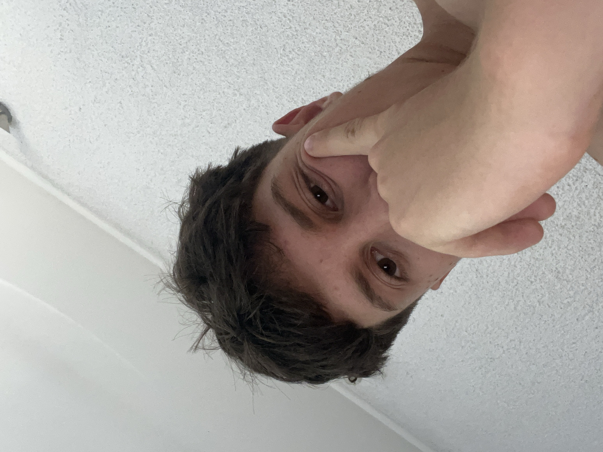 The image, taken in Zlatograd, Bulgaria, features a close-up, low-angle portrait of an individual, likely male, with fair skin and disheveled dark brown hair. The subject's right eye, which is brown, is visible and directed towards the viewer. His right index finger is pressed against the skin at the outer corner of his right eye, causing a slight indentation and crinkling of the skin. The rest of his right hand, with fingers curled, partially obscures the lower left portion of his face. The background consists of a plain, light-colored, textured surface, possibly a ceiling or wall, exhibiting a fine granular pattern. Subtle linear features, appearing as faint lines or shadows, are discernible in the background, suggesting architectural details. A small, dark, indistinct object is visible in the upper right background area.