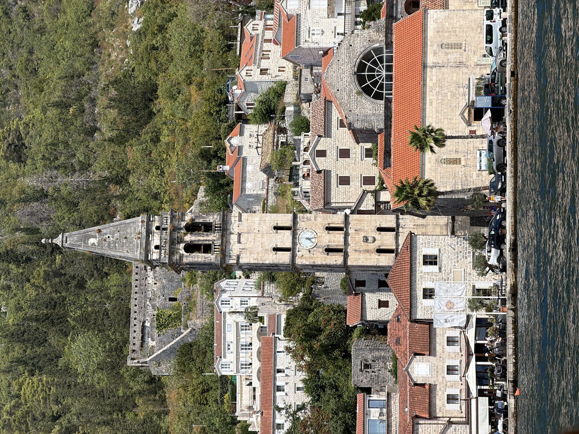 This is an outdoor, eye-level, vertical, medium shot of the waterfront of Perast, Montenegro, taken on a sunny day. The foreground is dominated by dark, rippling water. Behind the water, a stone embankment lines the shore, in front of which are several cars parked along a paved area. Beyond the paved area are buildings with red-tiled roofs. On the left, a restaurant with white awnings is visible, with patrons seated at tables outside. Further to the right, a large stone church with a rounded arched window occupies the middle ground. A prominent, tall, stone bell tower with a clock is situated to the left of the church, and appears to be the dominant architectural feature of the scene. Numerous stone buildings of varying sizes and architectural styles are built on the hillside behind the bell tower and church, ascending towards a forested slope covered in green trees. A section of stone fortifications is visible higher up on the hill. The sky is clear and blue.