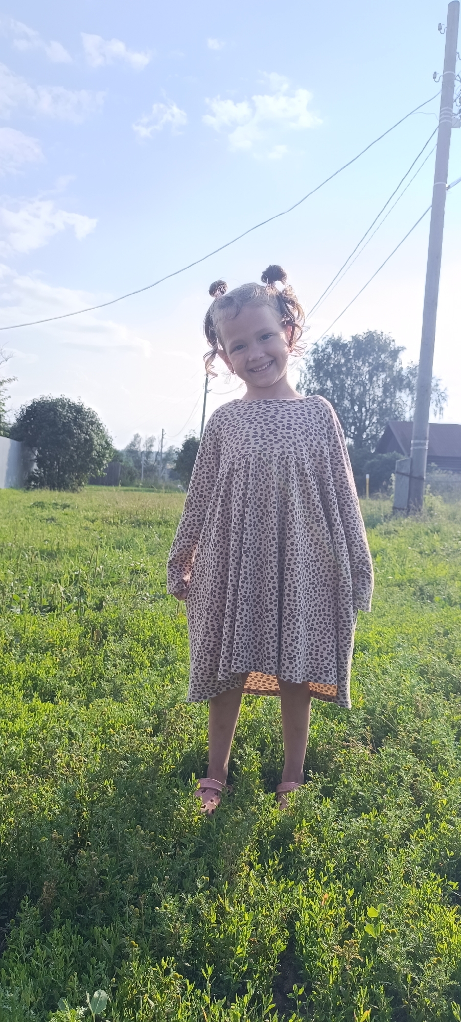 A young girl with curly hair styled in two buns stands in a grassy field, smiling at the camera. She is wearing a loose-fitting, long-sleeved dress with a leopard-print pattern and pink sandals. The background shows a power pole and wires, trees, and some houses in the distance. The sky is blue with scattered clouds, and the sun is casting a warm light. The location is identified as Karsha, Russia.