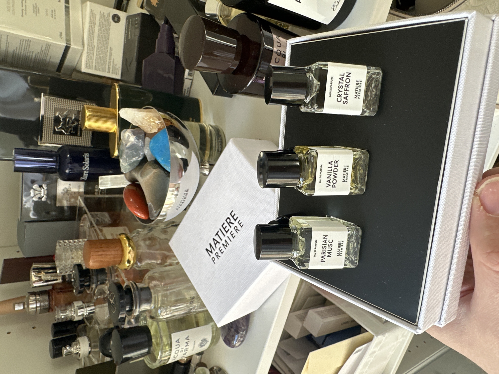 Here is a detailed description of the image:
The image captures a collection of perfumes and related items, arranged somewhat haphazardly on a shelf or surface, possibly in a personal setting like a vanity or storage area.
The central focus includes a white box labeled "MATIERE PREMIERE," along with a set of three sample-sized perfume bottles in a silver and black box. Each bottle has a black cap and is labeled: "CRYSTAL SAFFRON," "VANILLA POWDER," and "PARISIAN MUSC." All three bottles also have the "MATIERE PREMIERE" brand name on them.
Around the box and bottles, there's a collection of various other perfume bottles, some standing and some lying down. Brands visible include "ACQUA DI PARMA" and "Zure Solaris." There is also a glass container of stones.
The background is filled with additional boxes and bottles, suggesting a larger collection or storage space for beauty and fragrance products. The lighting is somewhat diffused. A portion of a hand is visible in the lower right.
No specific location in London, United Kingdom can be determined.