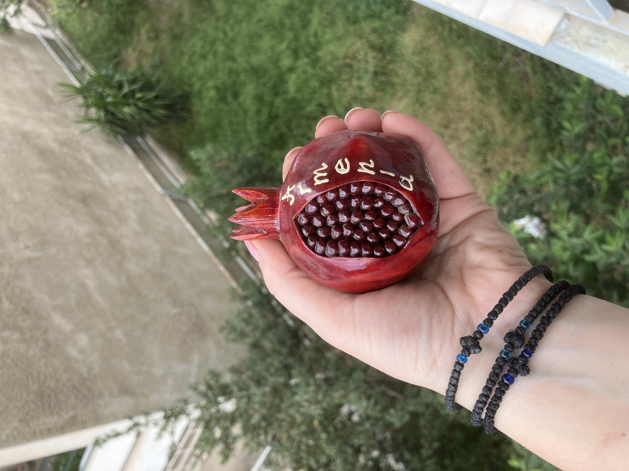 A high-angle, close-up shot features a person's hand holding a decorative pomegranate against a softly blurred outdoor background. The hand, with neatly manicured fingernails, wears three distinct black, braided bracelets, each accented with small blue beads and knotted details.
The central object is a meticulously crafted, dark red pomegranate ornament, possibly made of wood or ceramic, with a glossy finish. A section of the pomegranate is carved open, revealing a multitude of dark red, round "seeds" filling its interior. Prominently displayed on the exterior skin of the pomegranate, around the carved opening, are several light-colored, stylized characters resembling Armenian script.
The background is diffused and out of focus, showing vibrant green foliage that suggests trees or bushes, along with a light-colored, textured building wall or structure. This setting implies an outdoor location, possibly a balcony or an area overlooking a garden, under bright, natural daylight. The image focuses on the intricate details of the handmade pomegranate and the person's adornments, evoking a sense of appreciation for craftsmanship and cultural artistry.