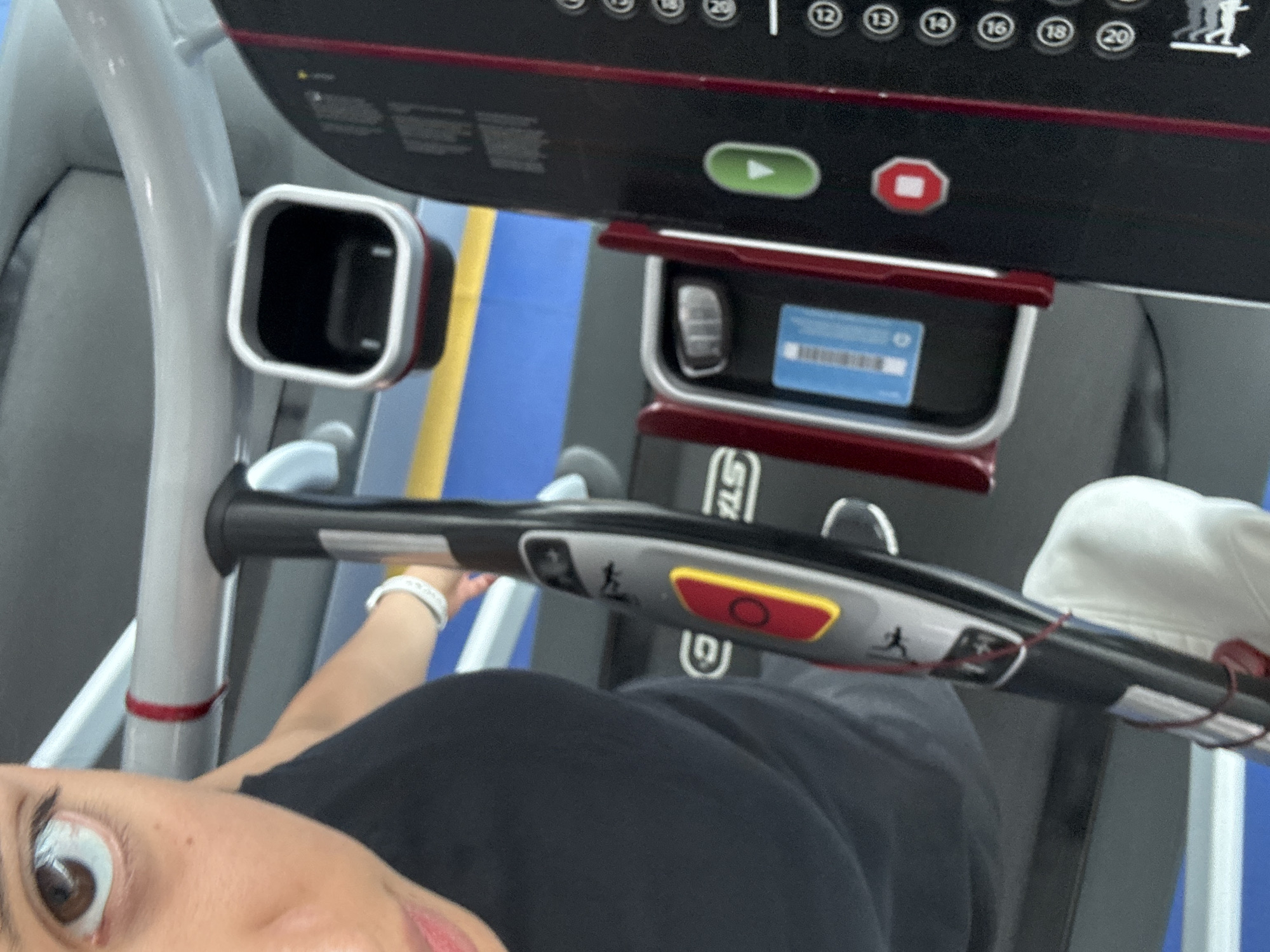 A selfie taken by a person at a fitness center in Heroica Puebla de Zaragoza, Mexico. The person, wearing a black shirt, is partially visible in the lower-left portion of the frame, with one eye and part of the face clearly seen. The focus is on the treadmill in front of them. The treadmill has a control panel with buttons, including a start button (green triangle) and a stop button (red square), as well as numbered speed settings. A device holder is visible below the panel. The treadmill's handles are in the foreground. The fitness center has blue and yellow striped walls and gray treadmill components. The person is wearing a white fitness tracker on their wrist.