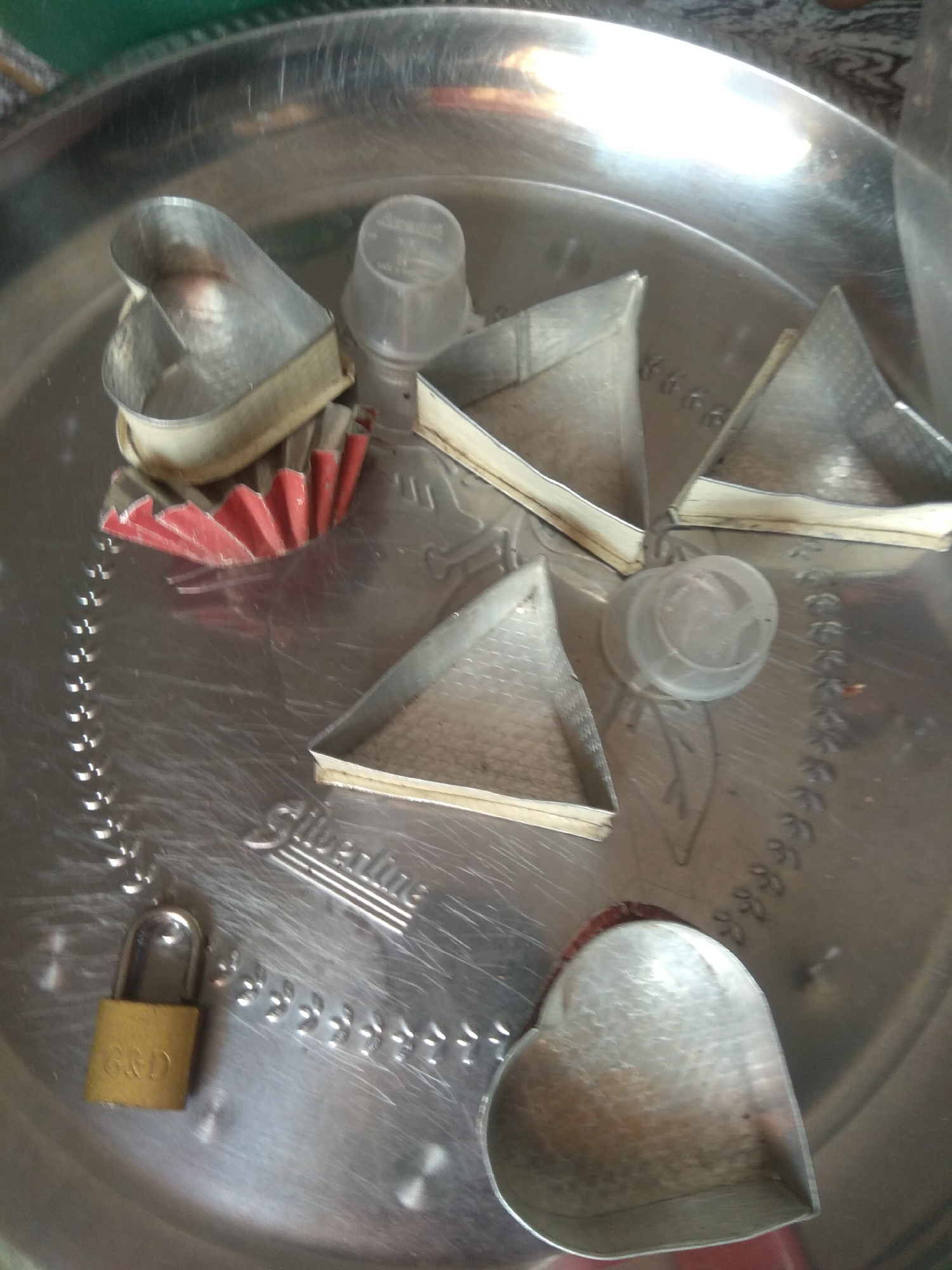 The image shows a collection of metal cookie cutters arranged on a round, metallic tray with a patterned rim. The cookie cutters include several heart shapes and triangular shapes. Some of the cutters have a white rim or are lined with red paper or plastic. There are also two clear plastic lids or caps on the tray. In the lower left corner, a small, gold-colored padlock with "G&D" inscribed on it is visible. The tray itself has embossed text that appears to read "Silverline". The lighting in the image is bright and reflective, creating glare on the metallic surfaces. The overall impression is of a collection of baking or crafting tools. This scene is not located in Jalingo, Nigeria, as no contextual information or visual cues suggest this location.