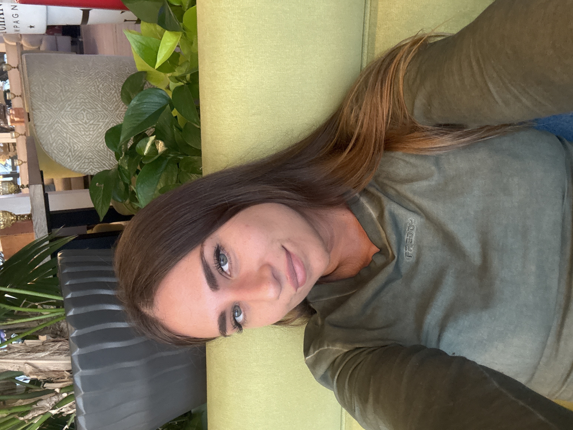 A young woman with long, brown hair and blue eyes is shown in a close-up, vertical, selfie-style shot. She is wearing a long-sleeved, olive-green t-shirt with the word "FREDDY" visible in a vertical line on the left chest area. She is lying down on a lime-green couch. The background includes indoor plants with lush green leaves and a glimpse of what appears to be shelving with decorative items. The lighting suggests it could be daytime. The woman's expression is relaxed and she appears to be smiling slightly. The setting is indoors, possibly a home or a cafe with a natural, relaxed ambiance.