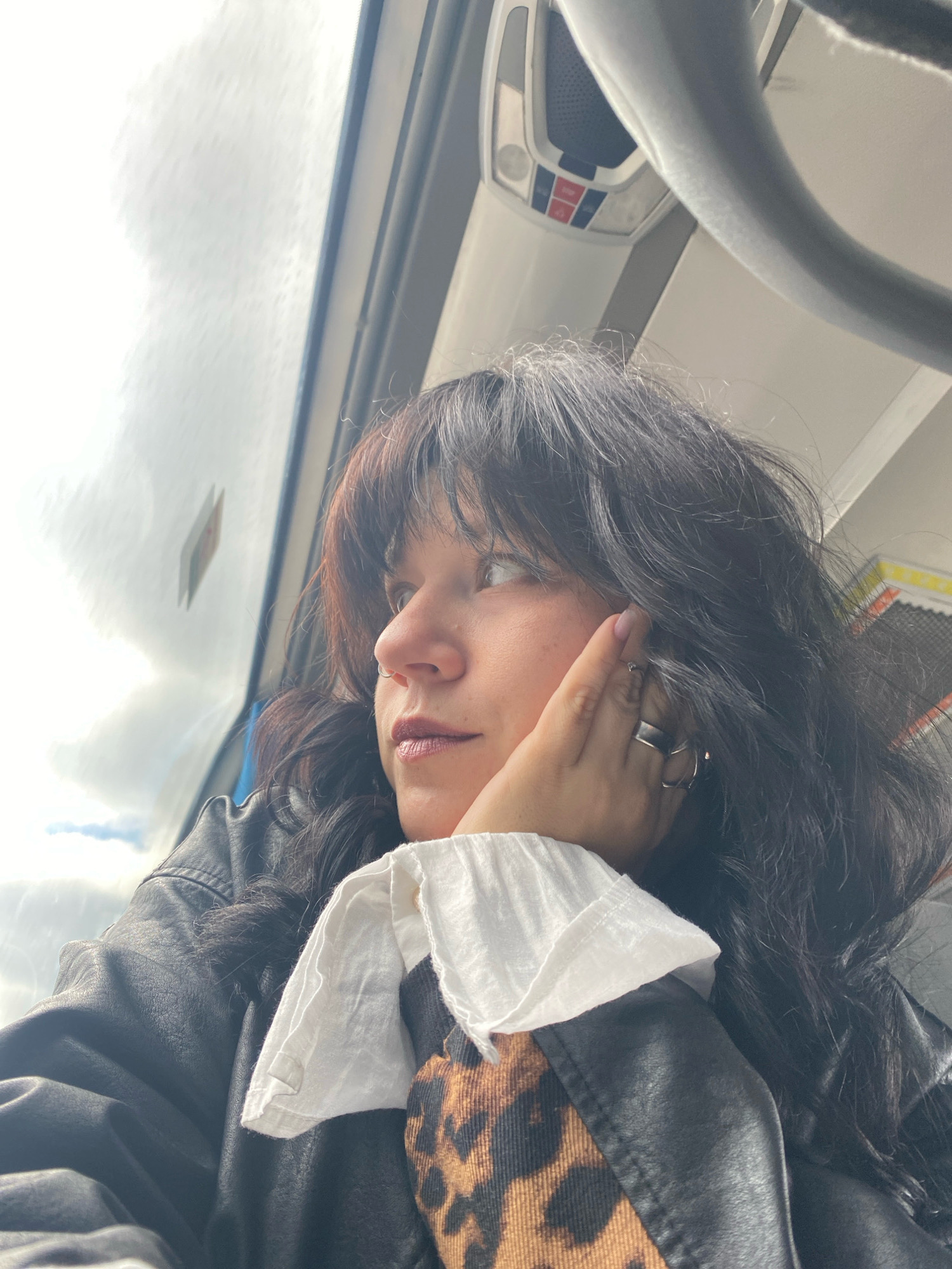 A young woman with dark, wavy hair is captured in a close-up, profile shot, looking out of a bus window. Her left hand is raised, with fingers resting gently on her cheek and temple, adorned with two silver rings. She is wearing a black leather jacket over a brown and black leopard-print garment, with the white collar of a shirt peeking out. The background suggests the interior of a bus, with a gray handle or railing visible above and to the right. Outside the window, the sky is overcast with diffused light and indistinct clouds. The scene is likely taking place on a bus in the city of Chaikovsky, Russia, as indicated by the provided context.