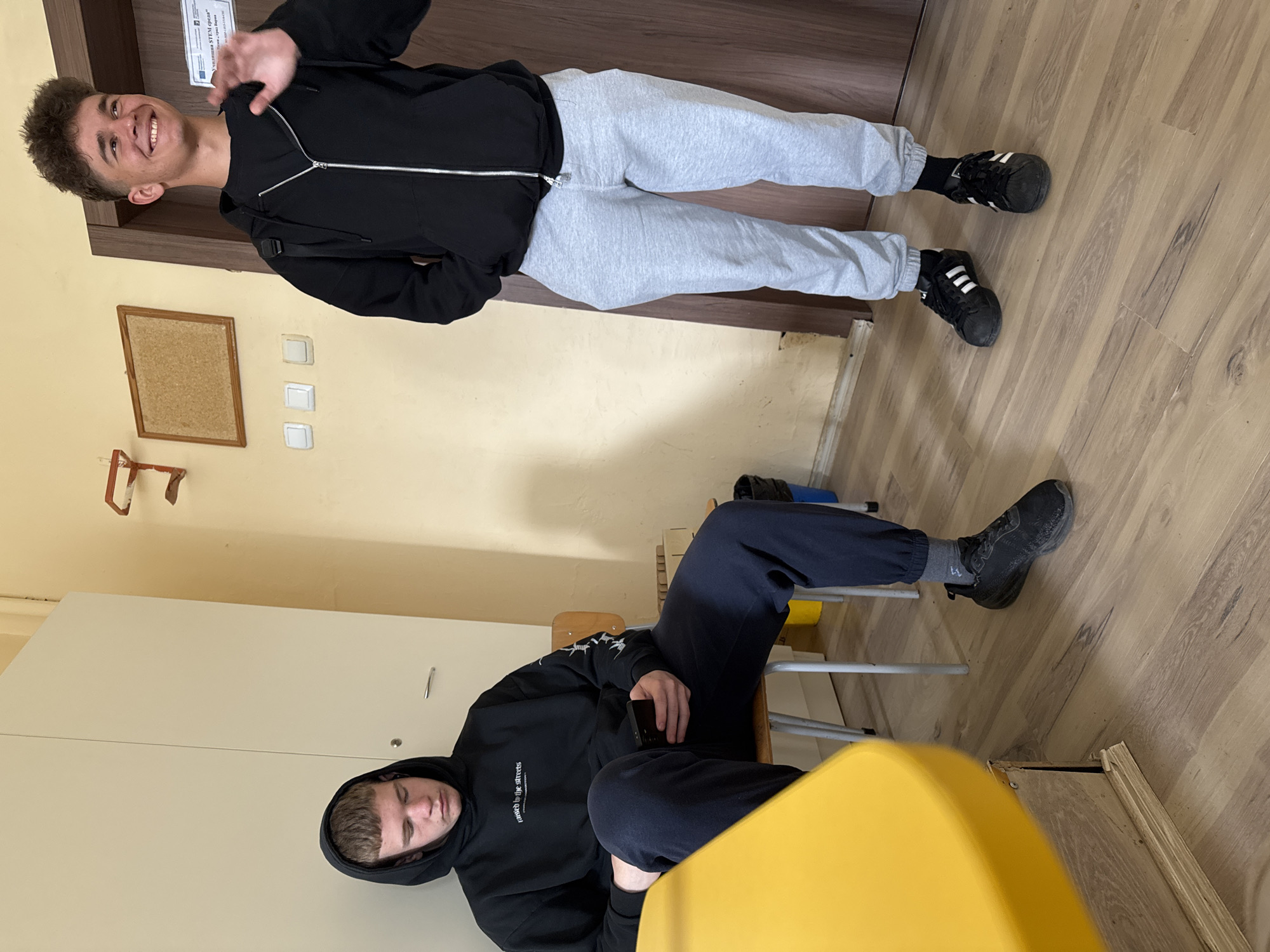 Two young men are in an indoor setting. The first man stands near a closed wooden door and light-colored wall. He wears a black zip-up hoodie, grey sweatpants, and black athletic shoes. He is smiling and holding up his left hand in a greeting gesture. The second man sits on a chair near a yellow table. He wears a black hooded sweatshirt and navy pants, and black athletic shoes. The room has light-colored walls and a light wood-look floor. A cork board and light switches are visible on the wall. The location appears to be an interior space in a building, not identifiable as being in Varna, Bulgaria.