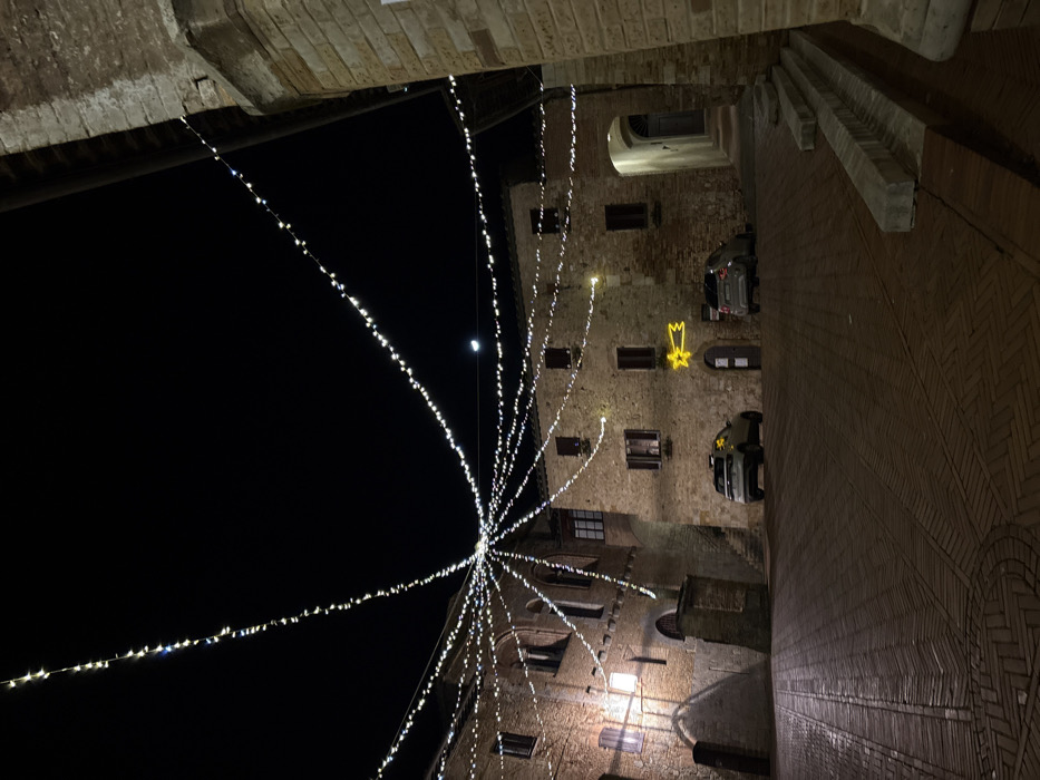 A nighttime view captures a building decorated with strings of lights radiating outward, forming a festive display. The building features brickwork and several windows, with a lit star hanging on one wall. Two cars are parked nearby. The sky is a dark contrast to the brightness of the lights and a faint crescent moon can be seen. The shot is taken from a low angle, looking upwards between the building and an archway.