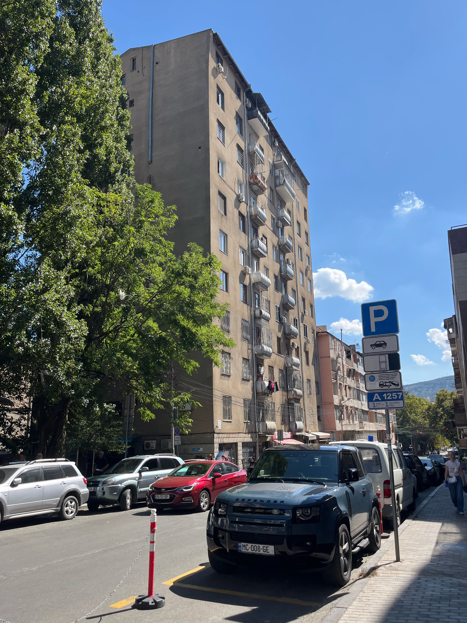 An eye-level, full shot captures a street scene in Tbilisi, Georgia on a sunny day, with a tall, light brown apartment building dominating the background. The building features numerous balconies with metal railings and some show clothes drying. A large, leafy green tree stands to the left of the frame, partially obscuring the building. Several cars are parked along the side of the street, including a silver SUV, a red sedan, a dark blue Land Rover Defender, and a white van. A pedestrian walks on the sidewalk to the right, and another person is partially visible in the distance. Parking signs with blue and white lettering are posted on a pole near the sidewalk. The sky is clear blue with a few scattered white clouds.
