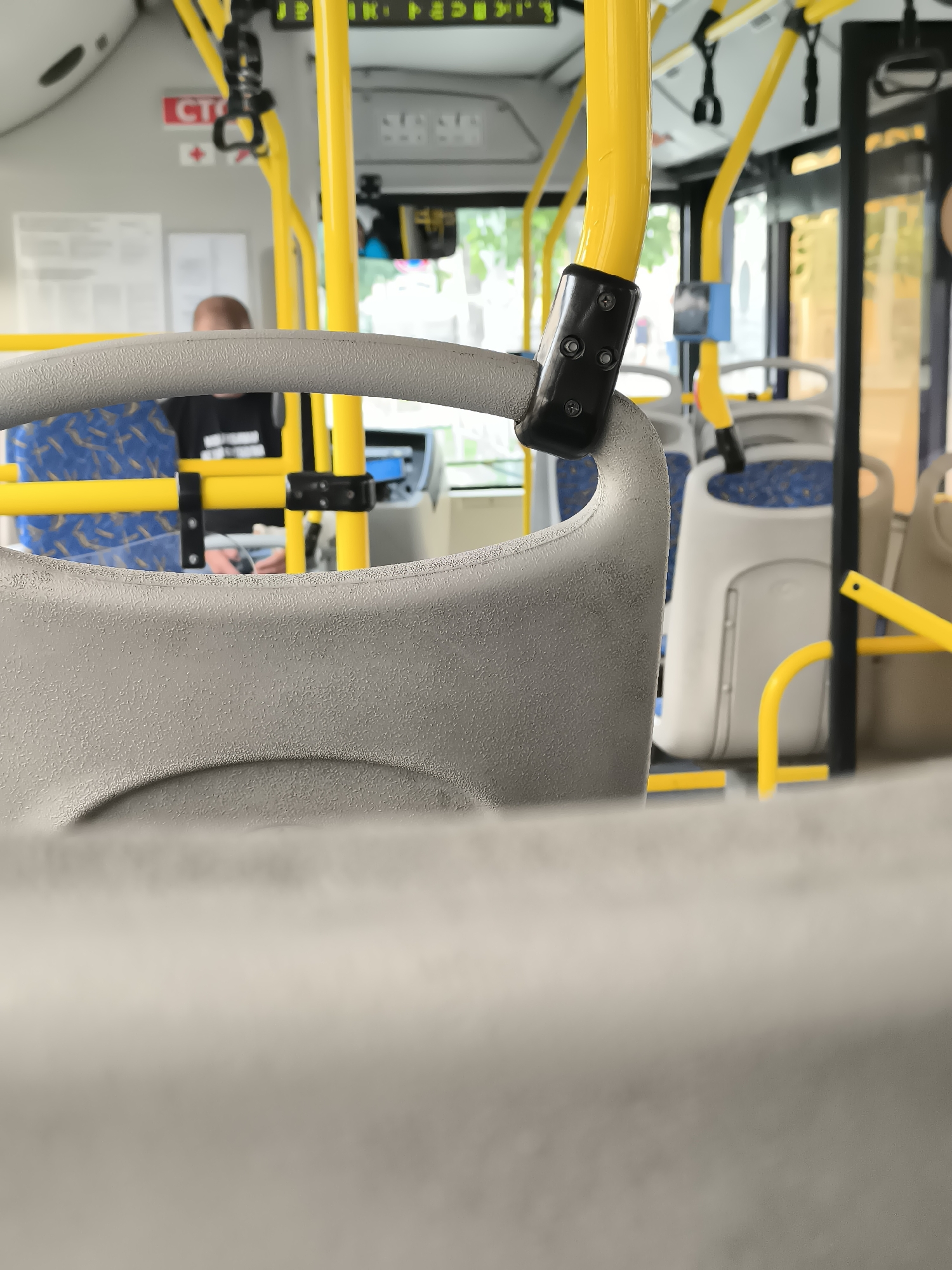 The image depicts the interior of a bus, likely in Astrakhan, Russia, given the context. The focus is on the gray, textured plastic of a bus seat's backrest, with a portion of a yellow handrail prominently visible in the foreground. Behind the seat, a person with a shaved head wearing a black t-shirt is seated. The bus has blue and yellow patterned seats and yellow vertical poles, typical of public transportation. The environment appears to be daytime, with light filtering in through the windows, suggesting the bus is either stopped or in motion.