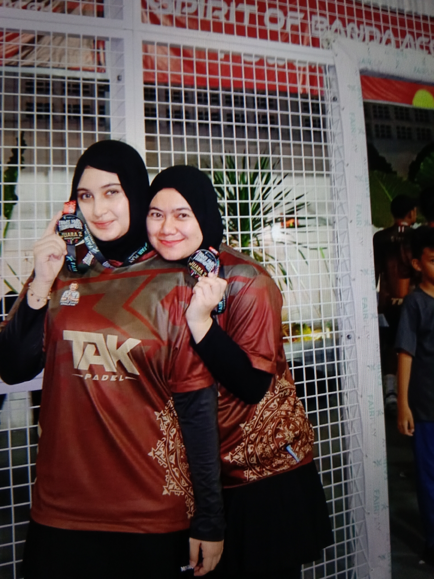 Two women, both wearing black hijabs, pose for a photograph in front of a white grid fence. The woman on the left has fair skin, dark eyebrows, and is holding a medal up to the camera. She is wearing a maroon long-sleeved shirt with a large "TAK PADEL" logo in gold lettering on the front. The woman on the right is also smiling and holding a medal. She is wearing a similar maroon shirt, but the sleeves are not visible. Both women are wearing black skirts. The background includes a grid structure, some greenery, and a banner with text that appears to be in Indonesian. A young boy in a dark t-shirt and blue pants is visible on the right side of the image, looking away from the camera. The overall scene appears to be an outdoor event or competition.