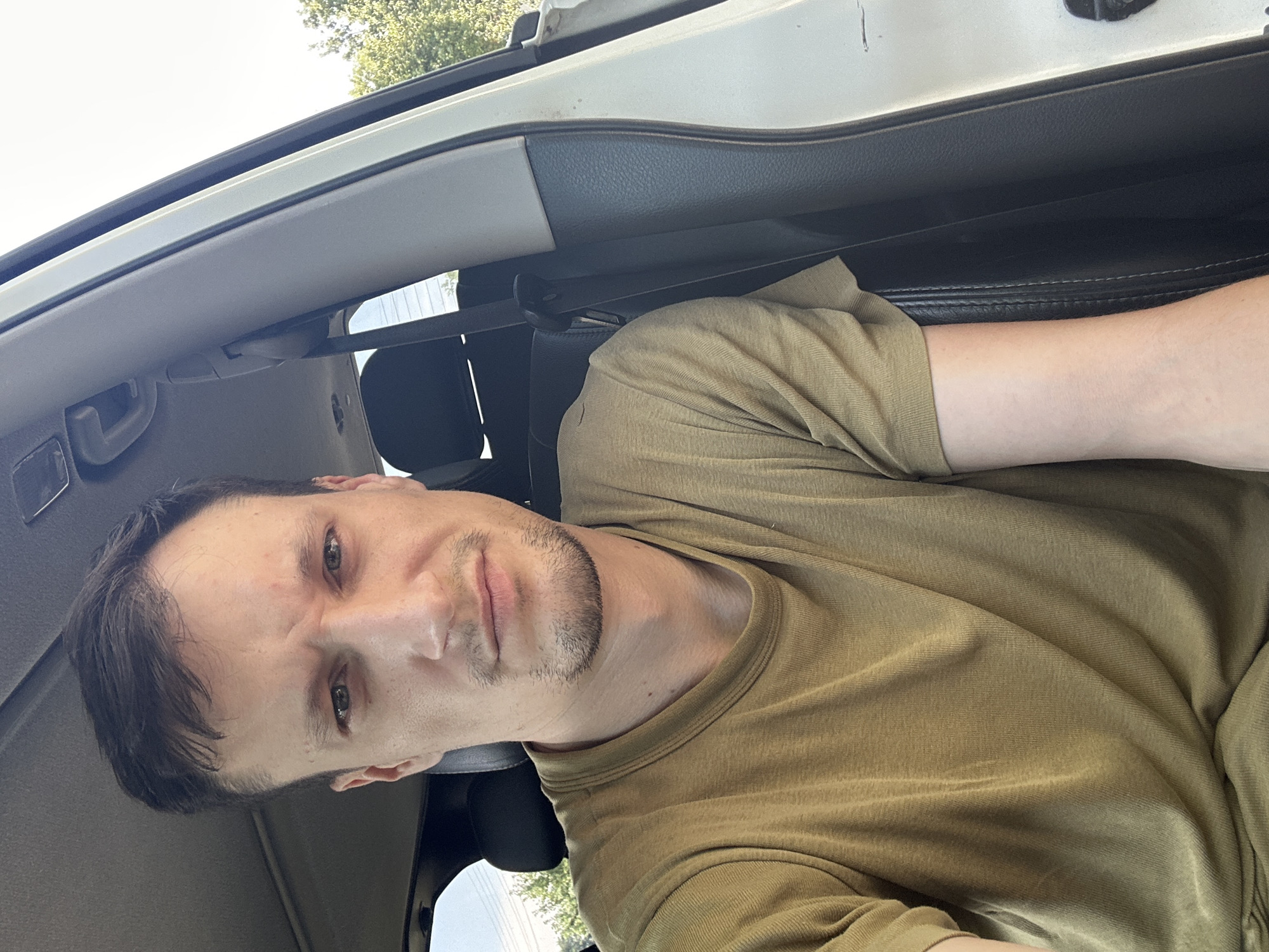 The image shows a close-up, chest-up, selfie-style photograph of a man sitting in the driver's seat of a vehicle. He has short, dark brown hair and a light beard. He is wearing a plain olive-green t-shirt. His expression is neutral to slightly concerned. The interior of the car is visible, including the dashboard and headrest of the passenger seat, as well as a portion of the door frame and exterior. The scene appears to be outdoors during daylight, with trees and a light sky visible through the car windows. The image is not specific to Makiv, Ukraine, as there are no identifying landmarks or contextual clues within the photograph to indicate this location.