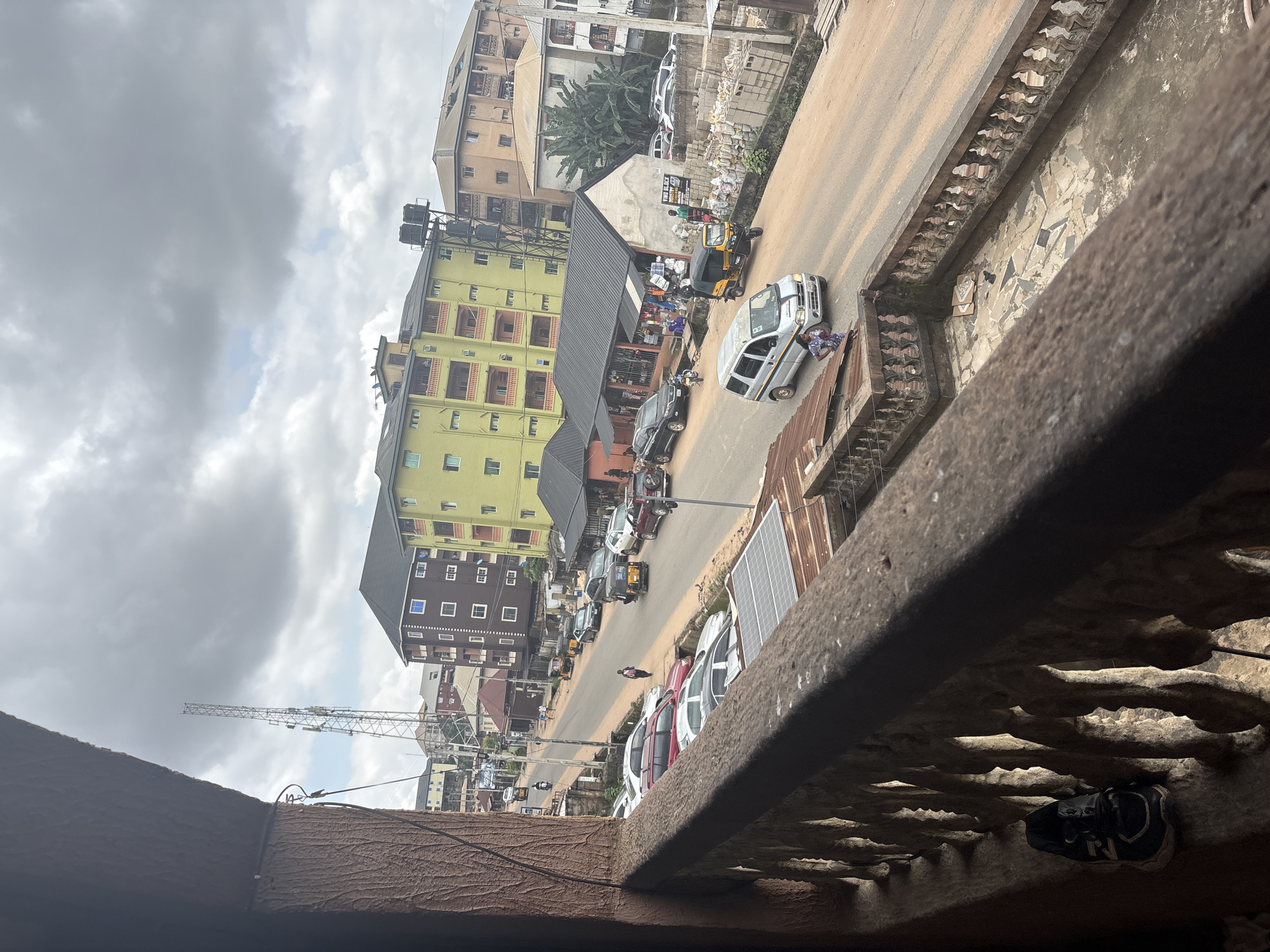 The image shows a street scene in Nkpor, Nigeria, viewed from a balcony. The street is lined with various buildings, including apartment complexes and smaller shops. Several vehicles are visible on the road, including cars, a minivan, and a three-wheeled auto-rickshaw. People are walking on the street and sidewalk, and some are standing near the shops. The sky is overcast with clouds. The balcony in the foreground is made of wood and concrete, and a pair of dark sneakers are visible on the railing. The overall impression is a busy urban environment on a cloudy day.