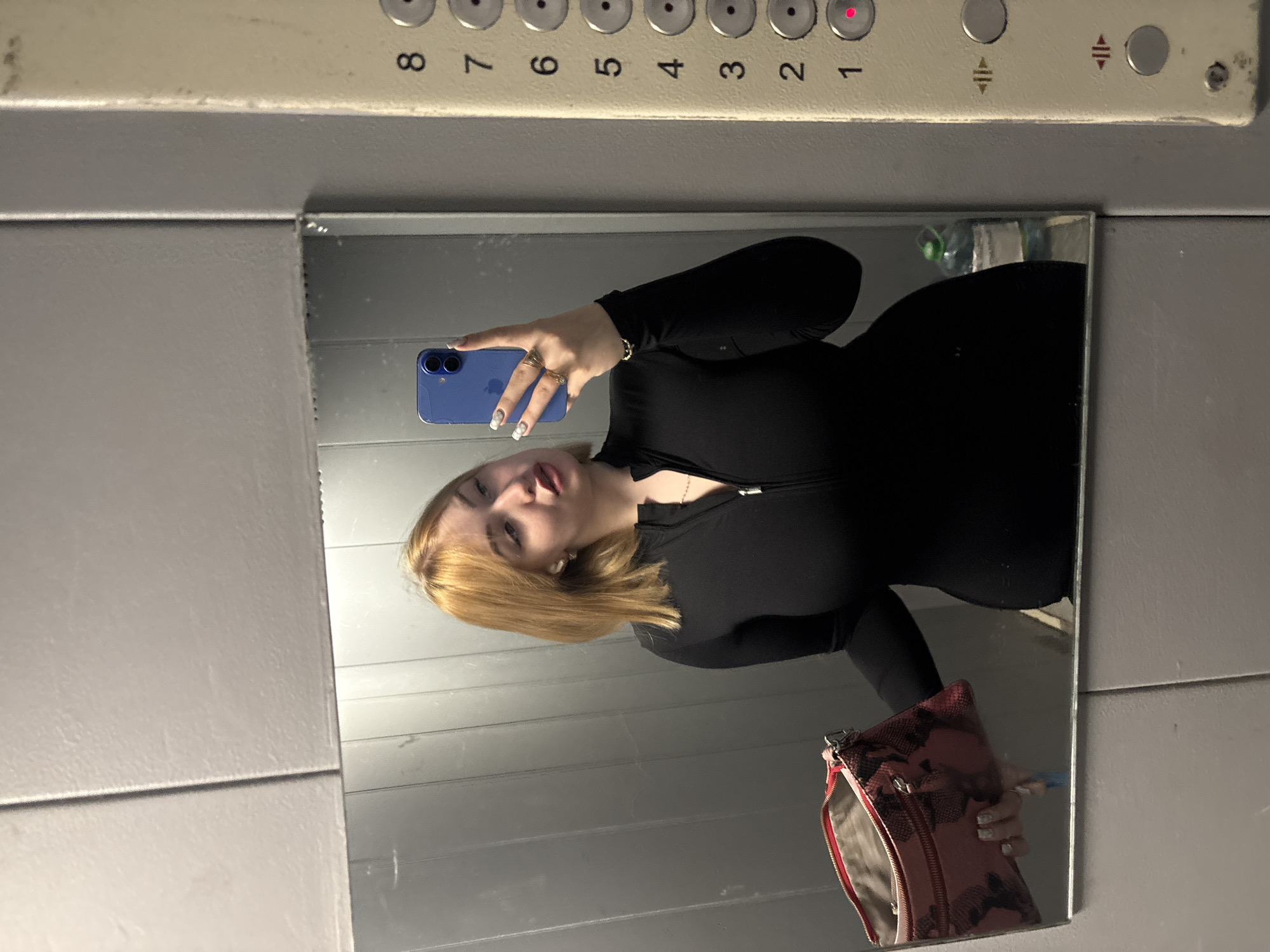 A young woman with blonde hair is taking a mirror selfie in an elevator. She is wearing a black, form-fitting, long-sleeved dress with a zippered neckline. She is holding a pink and black snake-print clutch in her left hand. In her right hand, she is holding a blue smartphone, which is pointed towards the mirror. She is wearing gold rings on her fingers and earrings in her ears. The elevator interior is grey with metal paneling. On the right side of the frame, an elevator control panel is visible with buttons numbered 1 through 8. The number 1 button has a red indicator light. A large bottle of water is visible in the reflection of the mirror behind the woman. The image is likely taken in an elevator in an apartment building.