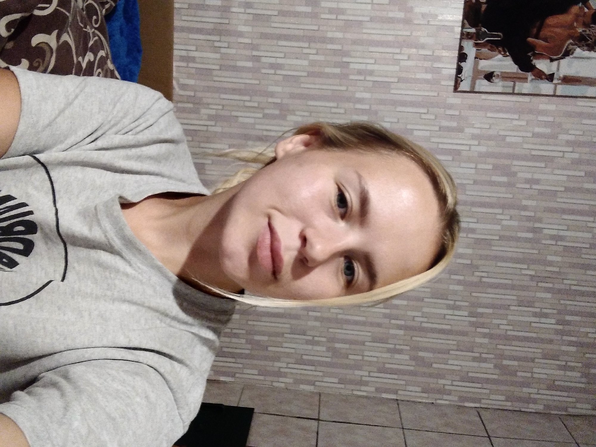 The image shows a young Caucasian woman with blonde hair and fair skin. She is wearing a light gray t-shirt. She is positioned in front of a wall with a vertically striped wallpaper. To the left of the woman, a framed picture is visible, depicting figures in a scene with wine. The overall environment appears to be indoors, possibly a residential setting. There is no identifiable information to confirm the location as Radisne, Ukraine.
