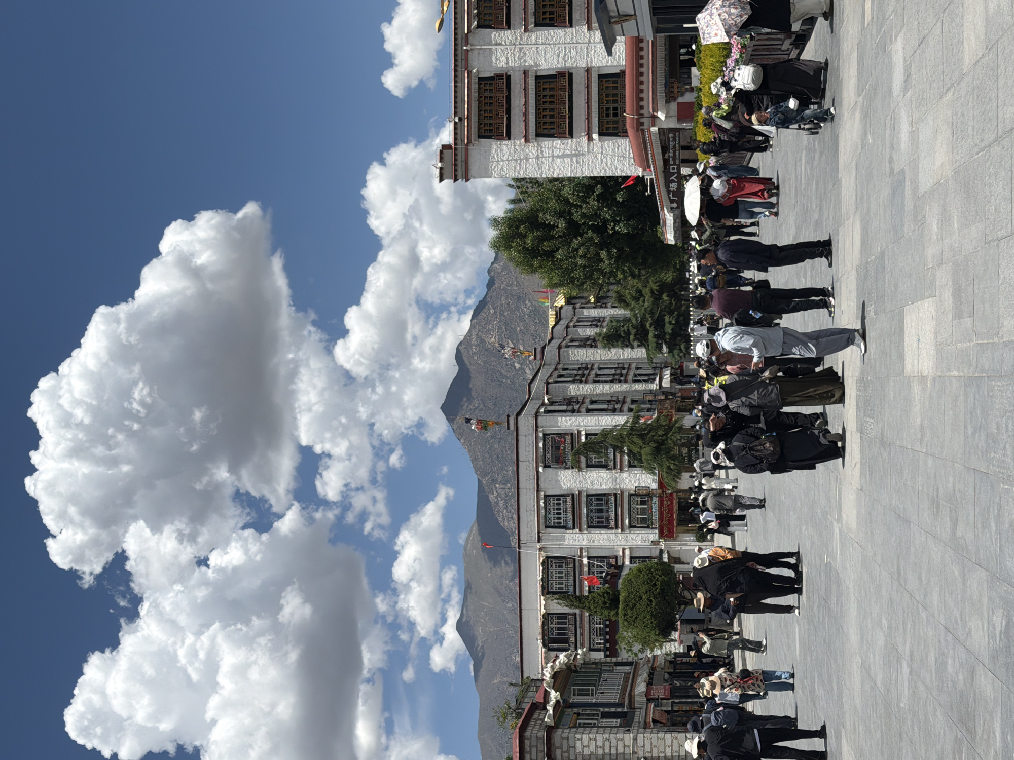 This image captures a vibrant public square or street scene in Chengguan District, China, under a clear, bright blue sky with large, fluffy white cumulus clouds. The strong sunlight and visible shadows suggest it is midday or early afternoon.
The scene is populated by numerous people walking, standing, and gathering across a wide, paved stone square. Individuals are dressed in a variety of casual clothing, with some carrying umbrellas, likely for sun protection. Their activities suggest everyday movement and congregation in a public space.
To the right of the square, a row of multi-story buildings showcases traditional Tibetan architectural elements, characterized by white-textured walls and dark-framed windows with intricate designs. Green trees are strategically placed near these structures, adding to the landscape. Colorful prayer flags are visible, draped from buildings and on the distant mountainside, serving as a prominent cultural cue.
In the background, a range of majestic, rugged mountains extends under the expansive sky. A red flag is seen flying from one of the buildings. A significant textual detail is a sign on a building that reads "广场入口" (Guǎngchǎng rùkǒu), which translates to "Square Entrance." The overall impression is one of a busy yet serene environment, reflecting the daily life and cultural richness of the area.