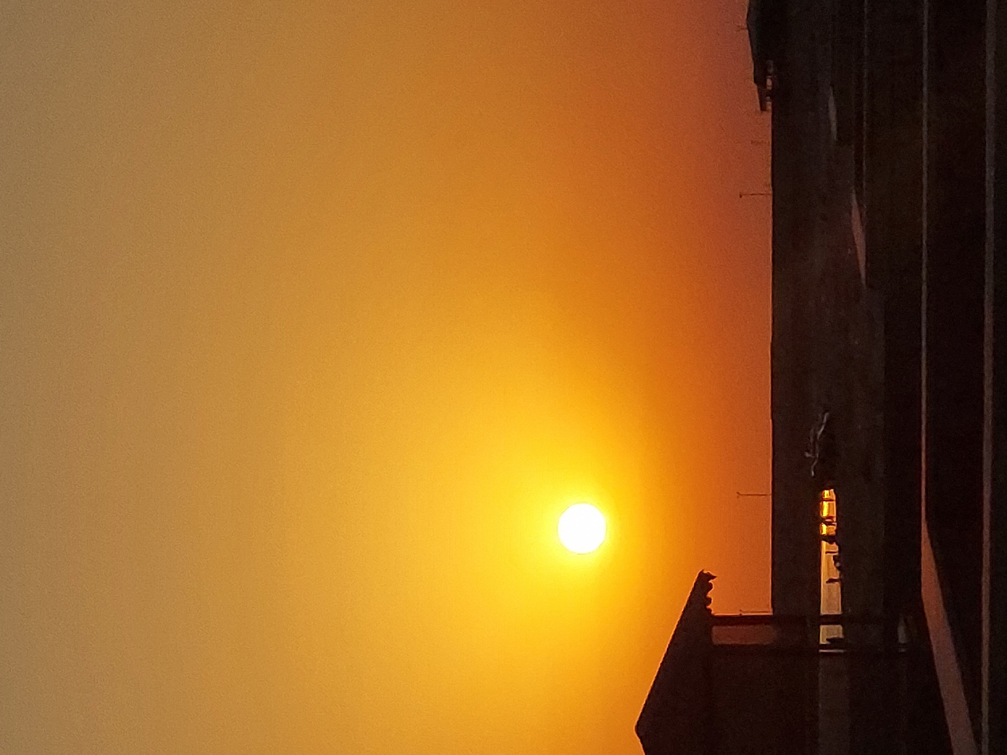 The image shows a sunset over the city of Pirallahi Raion, Azerbaijan. The sun is a bright orange disk in the center of the frame, casting a warm glow over the landscape. In the foreground, a silhouette of a building with a thatched roof can be seen, with several people standing near a body of water. In the distance, a flat horizon is visible, with a few structures and a street lamp. The overall mood of the image is peaceful and serene.