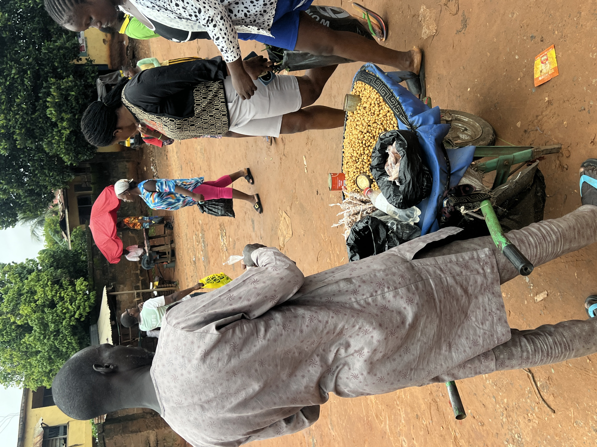 An outdoor market scene in Nkpor, Nigeria, features a man from the back view, wearing a light grey patterned tunic and trousers, holding a small clear packet. He stands next to a cart laden with yellow beans, some packaged in black bags. Behind him, several individuals are visible. A woman in a black top with gold lettering and white shorts walks away from the camera, while another woman in a white patterned top and blue shorts moves towards the foreground. In the background, people are gathered near a table under a red umbrella, and trees and buildings form the backdrop of the dusty street.