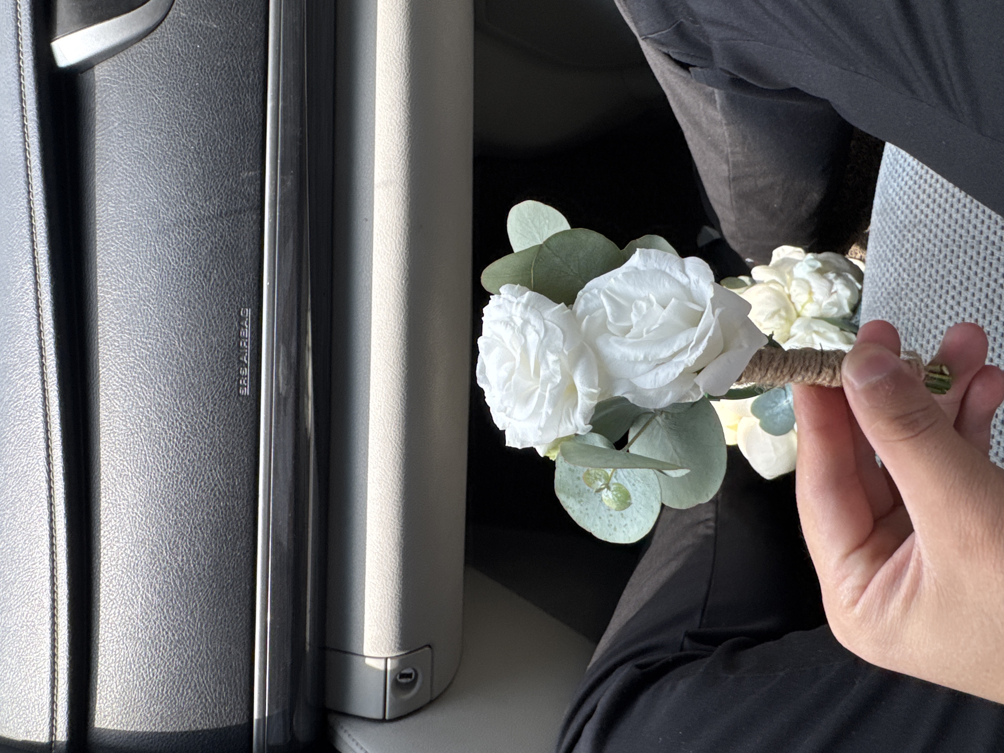 In Imeni Chapaeva, Kyrgyzstan, a person is holding a bouquet of artificial white roses and eucalyptus stems. The bouquet is tied with twine. The person is wearing black pants. The background appears to be the interior of a car, with the dashboard and a glove compartment visible. The lighting suggests it is daytime.