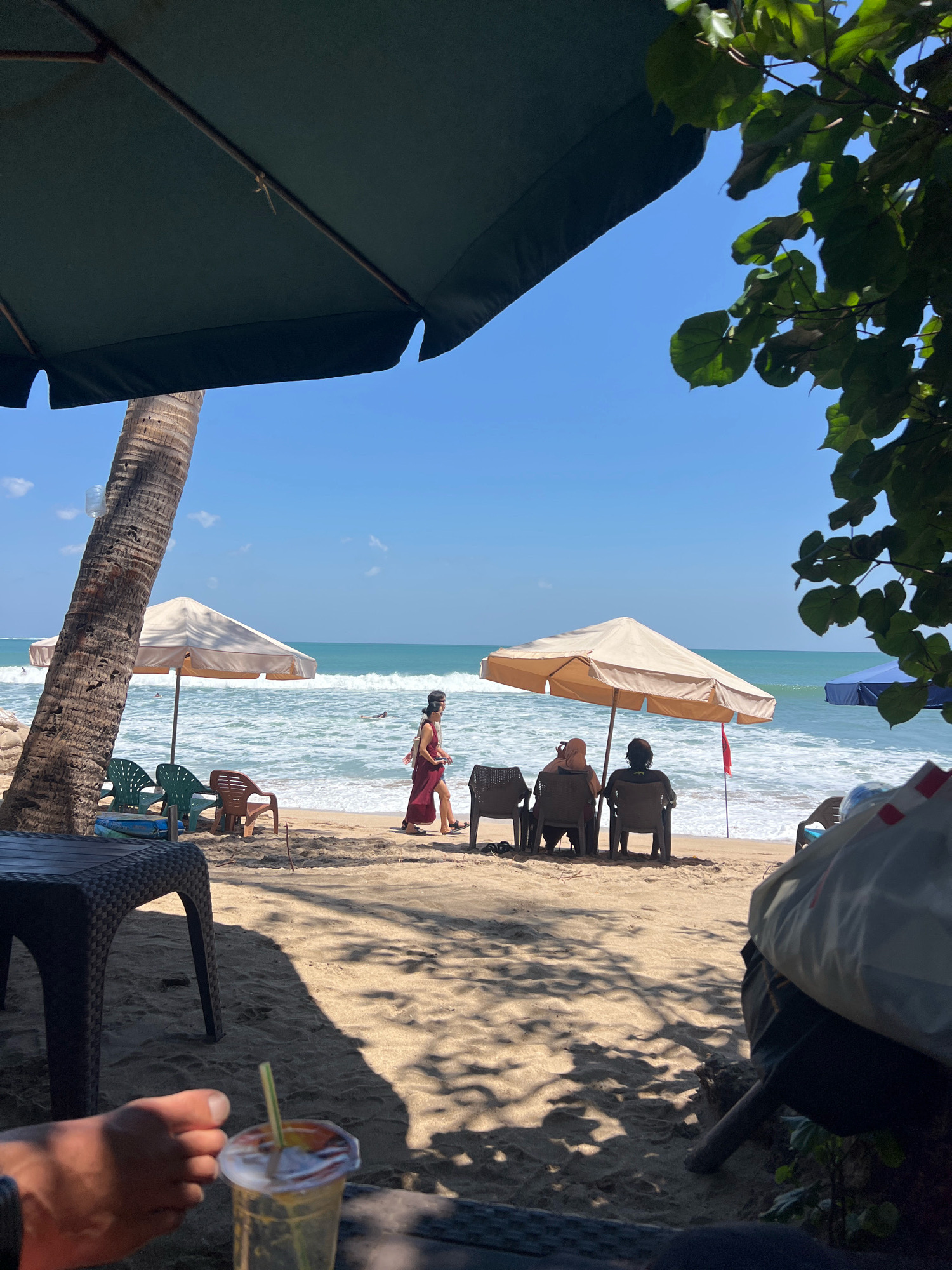 The image depicts a beach scene in Kuta, Indonesia, on a sunny day. In the foreground, a person's foot is visible next to a plastic cup with a straw, suggesting a relaxed setting. Two large, dark green beach umbrellas partially obscure the view of the sky and foliage. Beyond them, a tall palm tree stands to the left.
The sandy beach is occupied by several elements: a dark plastic table to the left, and several empty plastic chairs scattered around. Further back, two beige beach umbrellas provide shade for people sitting in chairs, facing the ocean. A single person, wearing a dark red dress, walks along the shoreline. The ocean is calm with gentle waves, and the sky is clear blue. In the background, another blue umbrella is visible further down the beach. The overall atmosphere is one of a leisurely day at the beach.