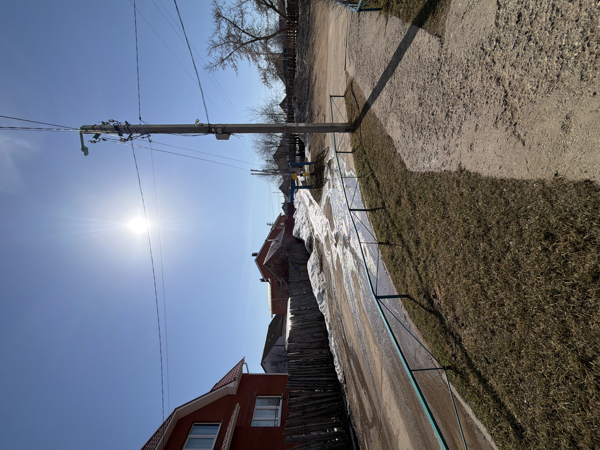 The image presents a sunlit street scene in what appears to be a residential area. A prominent feature is a utility pole extending into the sky, with numerous wires connected. The houses lining the street appear to be a mix of older and newer constructions, with varying roof types and materials. A wooden fence separates some of the properties. The road surface looks wet, possibly due to melting snow or ice. A green metal railing runs along the side of the road, separating it from a grassy area. The sky is a clear, bright blue. The perspective of the photograph gives a skewed sense of the orientation.