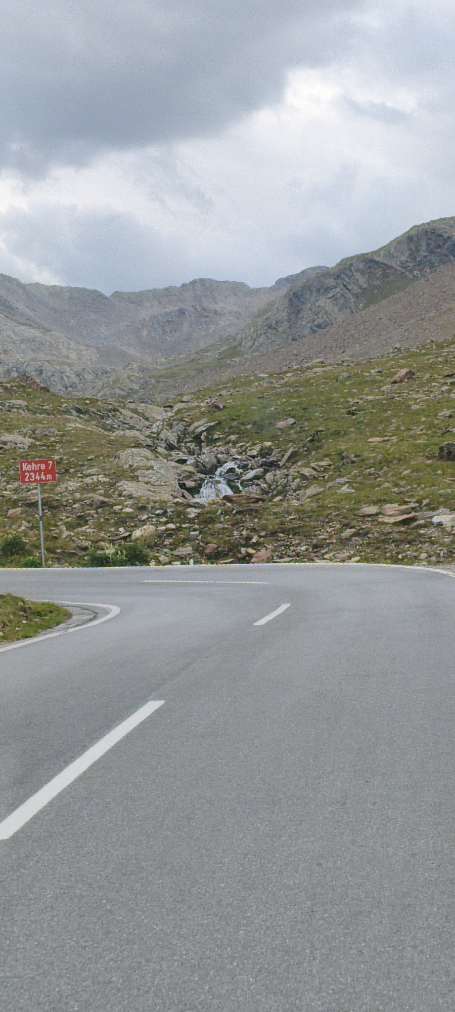 A winding asphalt road curves towards the viewer in the foreground, marked with white lines. In the middle ground, a red signpost with white text reads "Kehre 7 2344m". Beyond the road, a rocky mountainside is dotted with sparse green vegetation and a small waterfall cascades down the rocks. The background consists of rugged, gray mountains under a cloudy sky. The scene appears to be a mountain pass, likely in or near the Ötztal Alps, given the mention of "Kehre 7" which might refer to a hairpin turn on a mountain road.