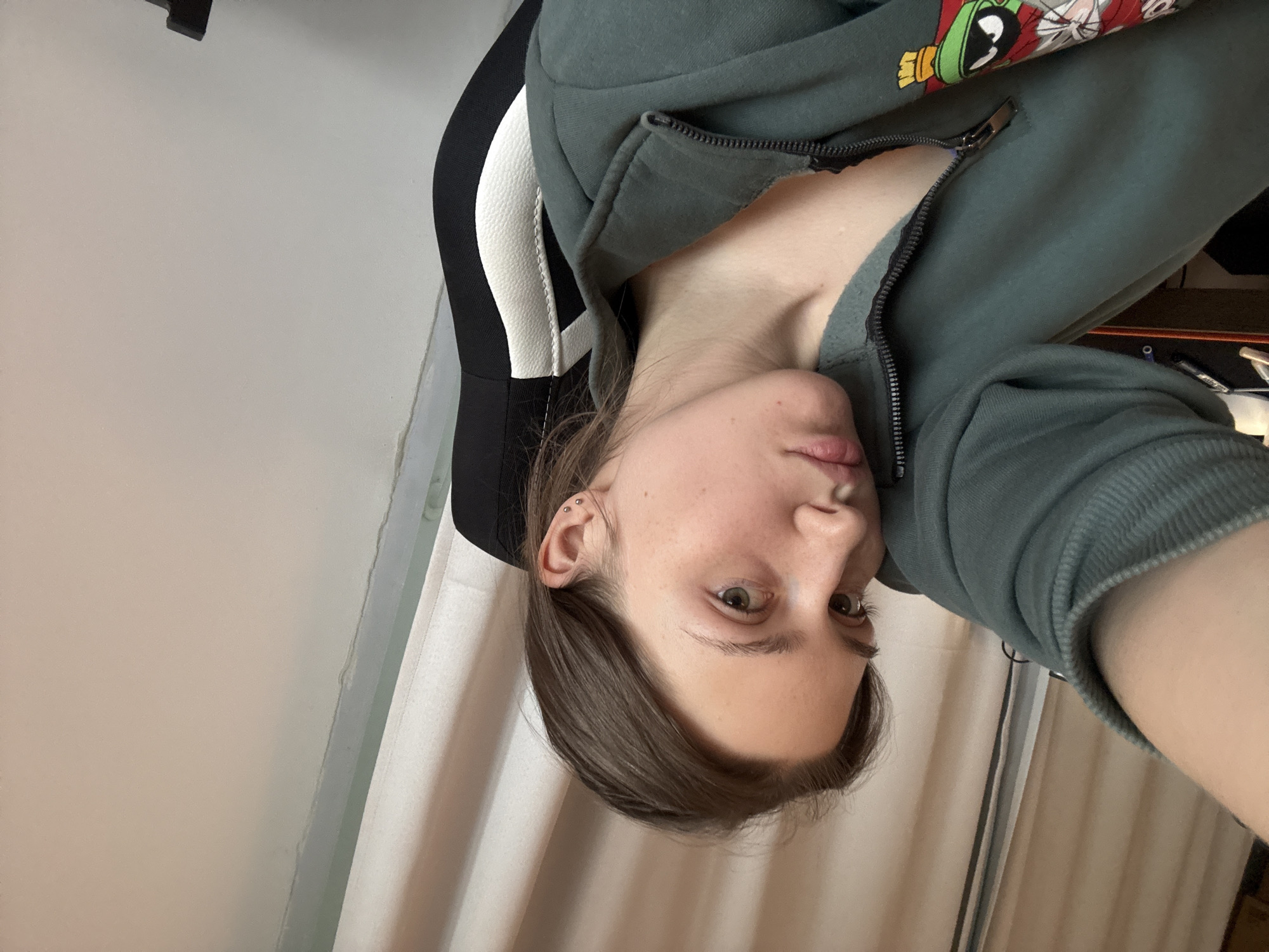 A person with fair skin, brown hair, and green eyes is shown in the image. The person is wearing a green zip-up hoodie with a graphic on the sleeve. The person is sitting in a black and white chair. The background consists of a white wall and a curtain. The location is not identifiable as Saint Petersburg, Russia, based on the image content alone.