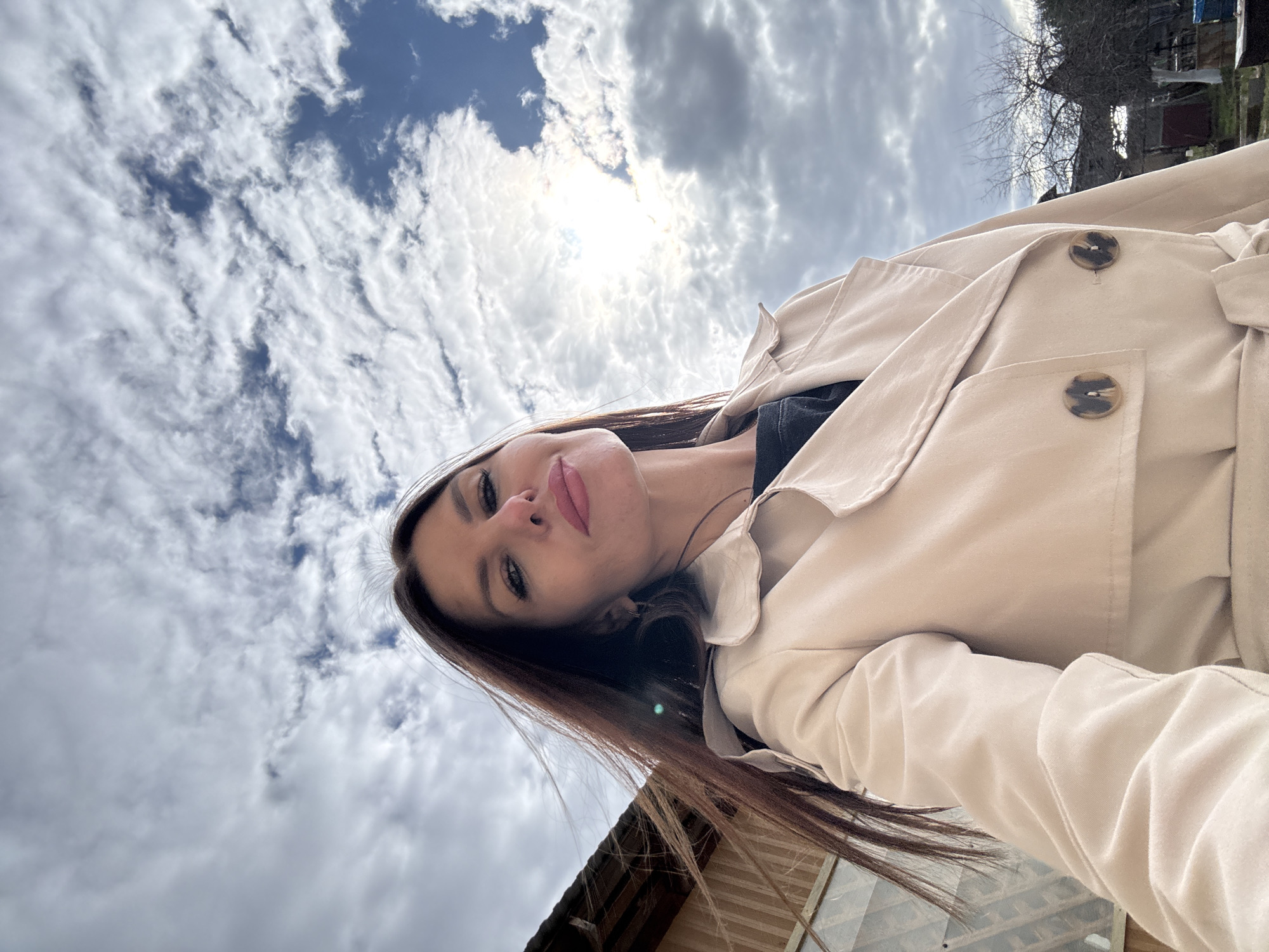 A photograph, oriented diagonally, shows a woman in the foreground. She has brown hair and is wearing a light beige trench coat. The background features a cloudy sky with the sun shining through. In the lower portion of the frame, a building with a window is visible. The location is Dubovaya, Russia.