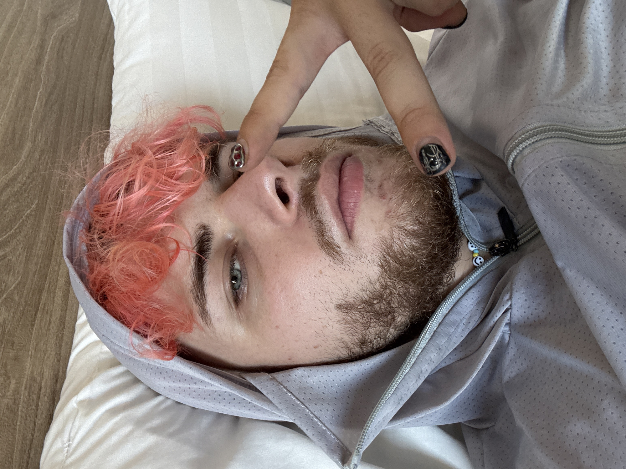 A person with pink hair, a beard, and light skin is lying on a white pillow. The person is wearing a gray hooded jacket and has dark nail polish with designs on their fingernails. The person is holding their hand up in a "peace" sign in front of their face. The background consists of a white pillow and a wooden surface. The location is not specified.