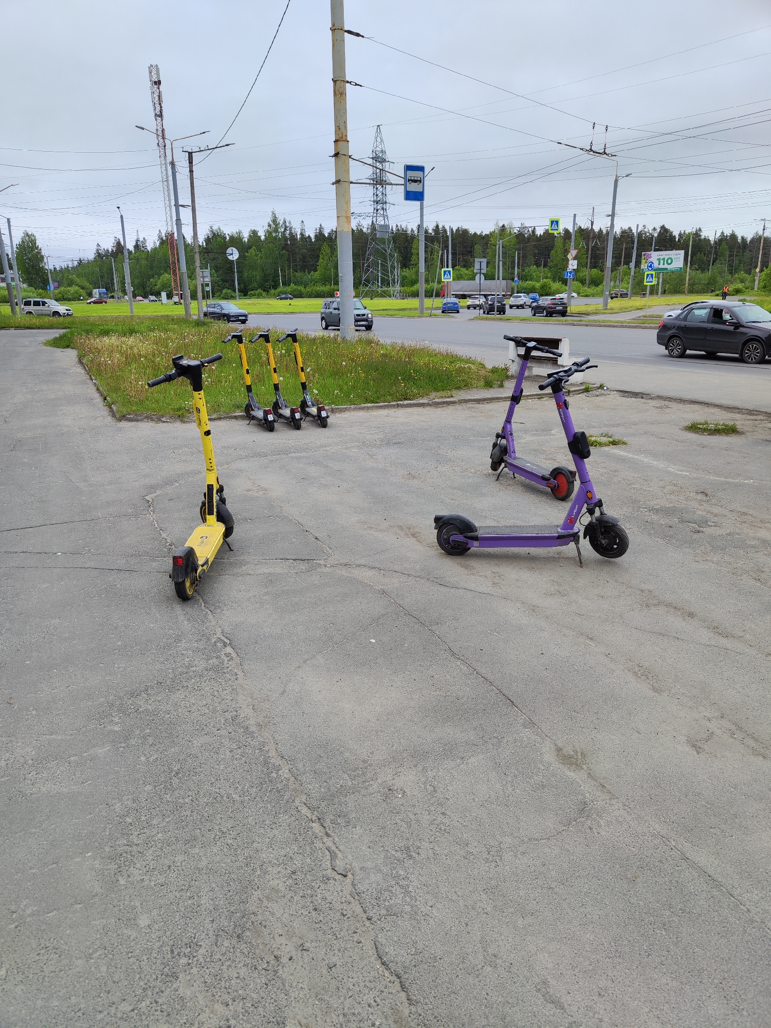 The image displays an outdoor scene featuring multiple electric scooters parked on an asphalt surface adjacent to a road, under an overcast sky. In the foreground, one yellow electric scooter, branded "Whoosh," stands facing right. Further back and to the left, three additional yellow "Whoosh" scooters are parked in a row. To the right, two purple electric scooters, branded "Юрент" (Yurent), are visible; the closer one faces left, with another parked behind it.
The scooters are situated on a grey asphalt area, which shows signs of cracks and wear. Adjacent to this paved section is a strip of grass with scattered dandelions. Beyond the grass, a multi-lane road is visible, with several vehicles, including a black sedan partially seen on the right, driving on the right side of the road. Other cars are in the distance.
The background is characterized by a dense line of trees, indicating a forested area bordering the road. The sky is uniformly grey, suggesting a cloudy or overcast day. Numerous utility poles with extensive wiring and power lines crisscross the upper portion of the frame. Several road signs are present: a blue square sign with a white bus icon (bus stop), yellow triangular signs with an "A" (pedestrian crossing ahead), and a large green sign displaying "110" with Cyrillic text "ЛЕТИ ЗА 110" (Fly for 110) and "до 900" (up to 900), which appears to be an advertisement. A person in an orange vest is faintly discernible in the far distance near the road. The visible Cyrillic text and road signs indicate a location in a Russian-speaking region.