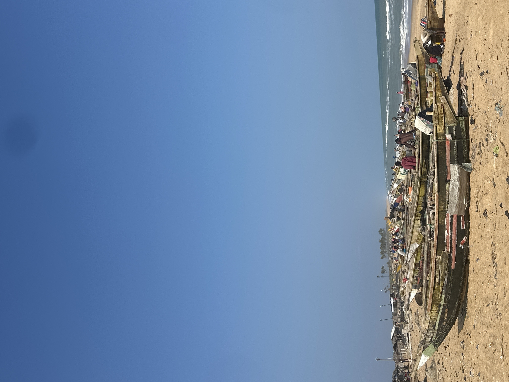 The image depicts a lively scene on a sandy beach, likely in Badagry, Nigeria. The primary subjects are numerous wooden fishing boats lined up on the shore, with several people actively engaged around them. The boats, varying in size and condition, are packed closely together, suggesting a busy fishing community.
People are seen tending to the boats, some appearing to be involved in repairing or loading them, while others are resting or socializing. The presence of various items and materials around the boats indicates ongoing activity, possibly preparation for or the aftermath of a fishing expedition. The overall atmosphere appears industrious and communal.
The setting is a beach with a clear blue sky dominating the upper left portion of the image, suggesting fair weather and possibly mid-morning or afternoon. The ocean is visible in the background, with gentle waves lapping at the shore. The sand is light brown and appears to be littered with some debris, typical of a working beach. In the distance, a line of palm trees can be seen, further reinforcing the tropical coastal location.
There is no visible text in the image. The lighting and the activity suggest a time of day when people would be actively working on the beach. The scene evokes a sense of daily life and livelihood in a coastal community.