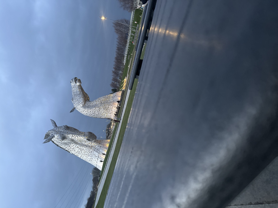 The scene depicts The Kelpies, two colossal equine head sculptures, located in The Helix Park, Falkirk, United Kingdom, at dusk or early evening. The two metallic sculptures, constructed from numerous steel plates, rise prominently from a landscaped green area. The sculpture on the left faces partially towards the viewer, exhibiting a more upright neck, while the one on the right faces right, with its neck in a more downward, arching posture. Both sculptures are illuminated from their bases with warm light.
The sky above is overcast and dark grey-blue, indicative of twilight. The ground consists of manicured grassy lawns and paved pathways. Several small, indistinct human figures are visible near the bases of both sculptures, appearing to be observing or walking around them. One figure stands near the left sculpture's base, and multiple figures are discernible near the right sculpture's base.
In the mid-ground, a line of slender, bare-branched deciduous trees is visible, extending towards the right. A tall street light with an illuminated fixture stands further right. The immediate foreground is dominated by a dark, flat, and horizontally textured surface, likely the top of an outdoor table or bench, which occupies approximately the lower half of the image. Reflections of ambient light are visible on this surface. The perspective is a low-angle shot looking up towards the sculptures.