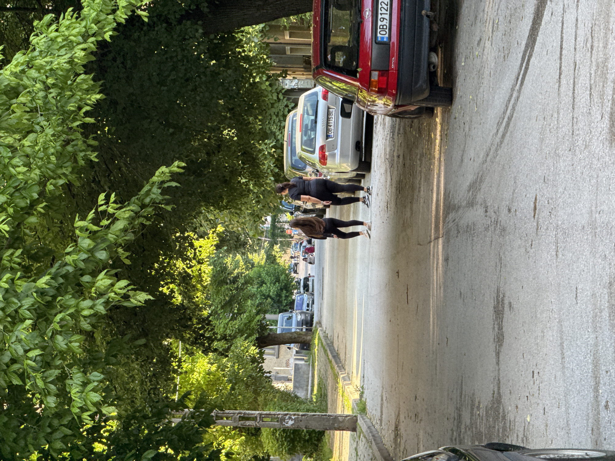 The image depicts a residential street in Lovech, Bulgaria, partially shaded by trees. Two women walk away from the camera, down the street. The street is lined with parked cars, including a red Opel with license plate OB 9122 and a silver Audi with license plate OB 4115 BE. Buildings are visible in the background, and a concrete post stands on the left side of the road.