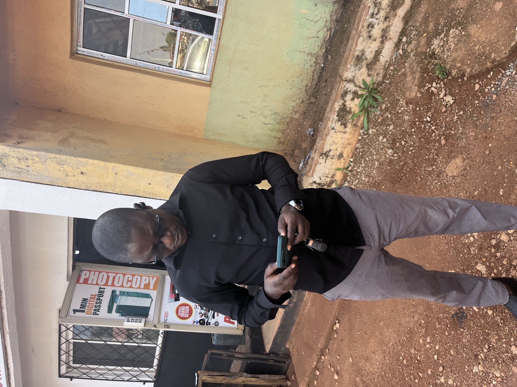 A man in a black button-up shirt and grey trousers stands on a reddish-brown dirt path in front of a building. He is looking down at his phone, which he holds in his hands. A set of keys hangs from his right hand. Behind him, a storefront displays signs for "1 MIN DIGITAL PASSPORT" and "PHOTOCOPY." The building has a yellow and light green facade, with a window above a textured wall. The ground is covered with loose rocks and sparse vegetation. The scene appears to be outdoors in an urban or semi-urban environment, possibly Gura Top, Nigeria.