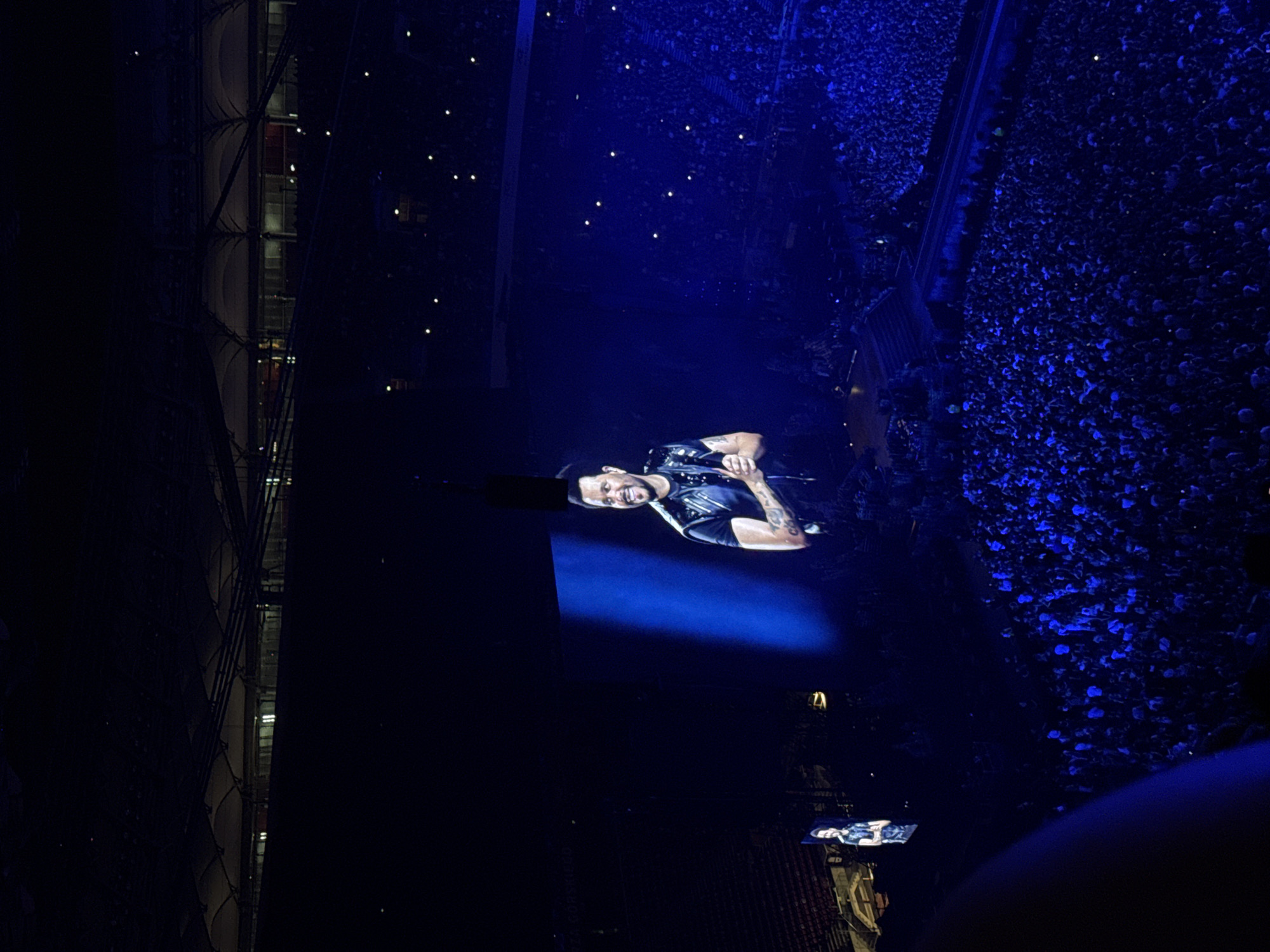 The image depicts a large concert venue at night, likely an indoor stadium, in Warsaw, Poland, illuminated by blue stage lighting and the glow of mobile phones in the audience. A prominent large screen displays a close-up of a smiling male artist wearing a dark vest over a t-shirt, with visible tattoos on his arm. To the left of the main screen, a smaller screen shows a full-body image of the same artist. Thousands of spectators are visible, packed into the seating areas and the arena floor, many holding up their phones, creating a sea of small lights. The architecture of the stadium roof is visible in the upper portion of the image, with rows of seating receding into the darkness. The overall atmosphere is that of a live musical performance with a significant audience.