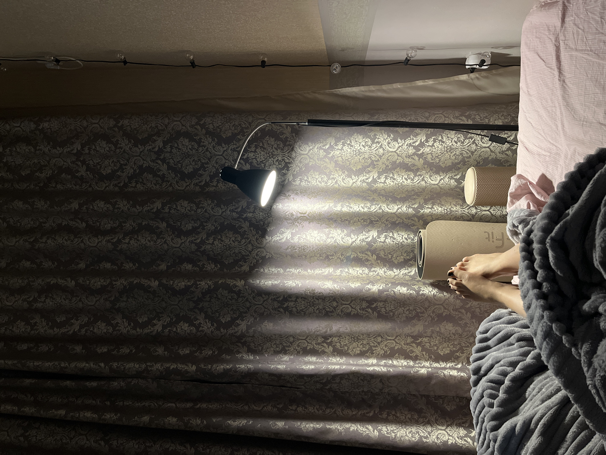 The image shows a room interior at night. The room's wallpaper is a patterned gray and gold. A black desk lamp illuminates the center of the frame. A yoga mat lies on the bed, next to a person's feet, which are partially covered by a gray blanket. A pink blanket is also visible. String lights are mounted on the ceiling. There is no explicit indication of this image being located in Sochi, Russia.